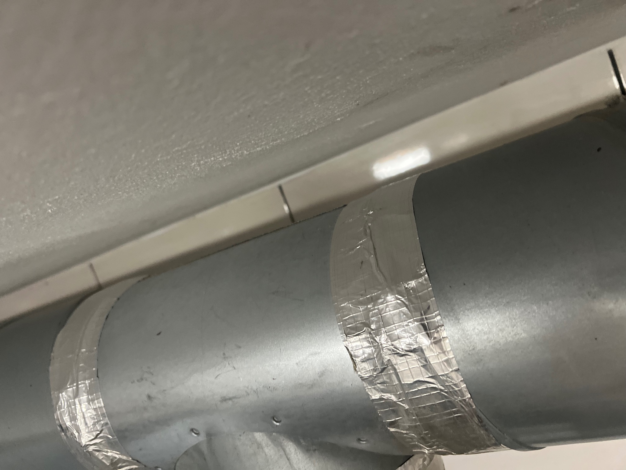 Metalowa rura wentylacyjna pod sufitem, owinięta srebrną taśmą naprawczą w dwóch miejscach.
