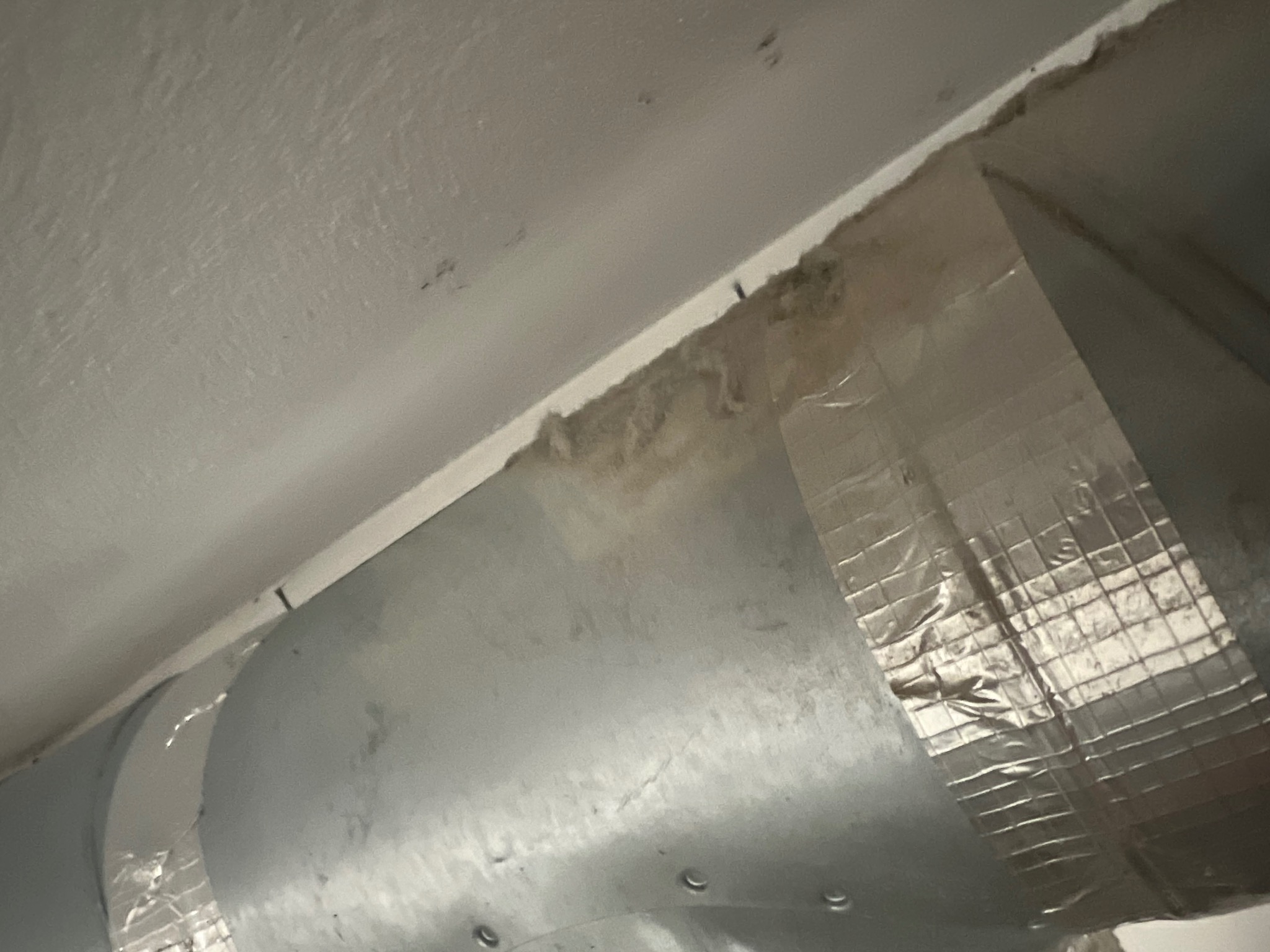 Zanieczyszczony kanał wentylacyjny przymocowany srebrną taśmą do sufitu, widoczne nagromadzenie kurzu i zabrudzeń wzdłuż krawędzi.