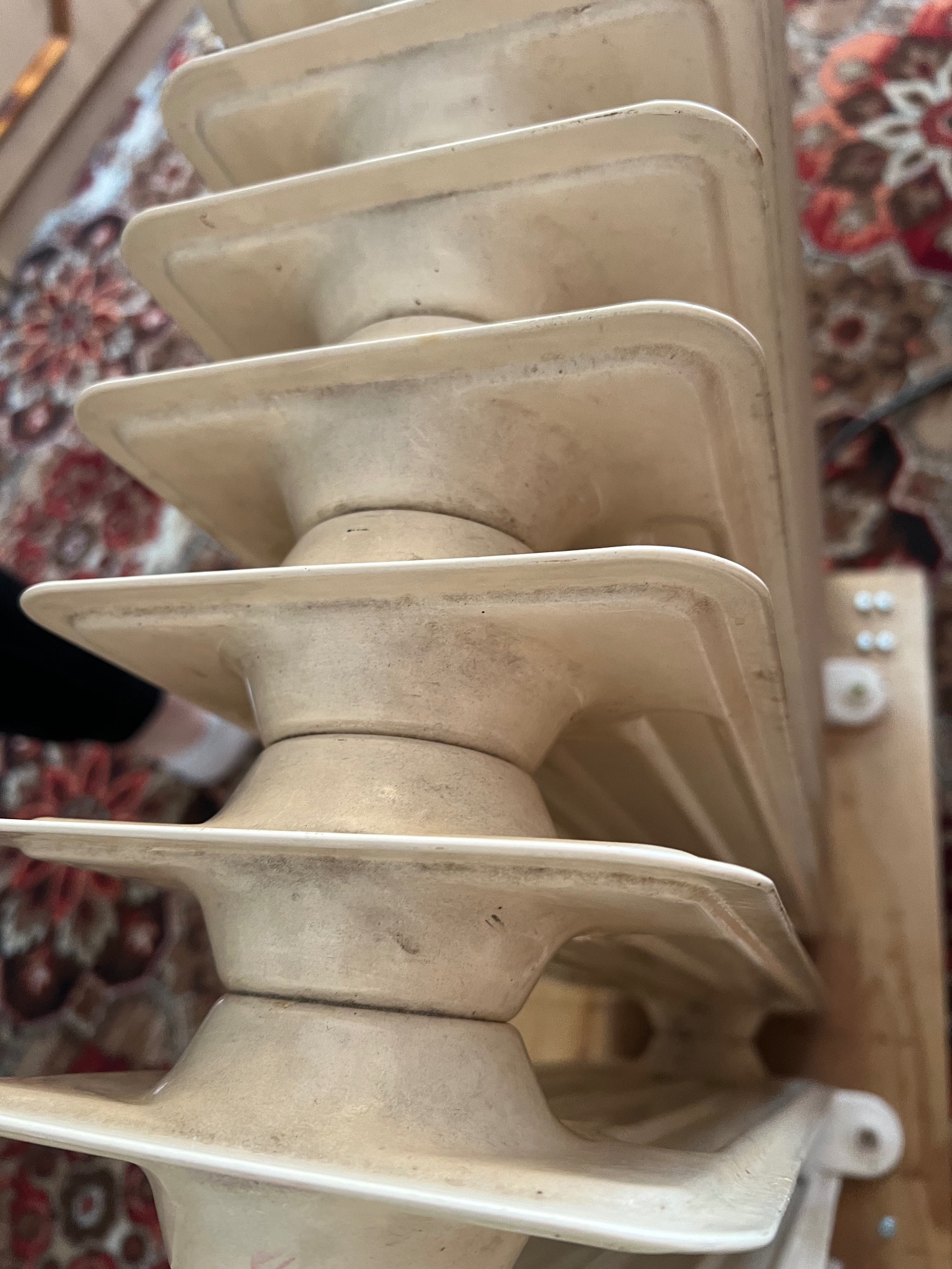 Żeliwny, kremowy grzejnik żeberkowy, wymagający odświeżenia, na tle wzorzystego dywanu w Olszowie. Widoczne drobne zabrudzenia i ślady rdzy.