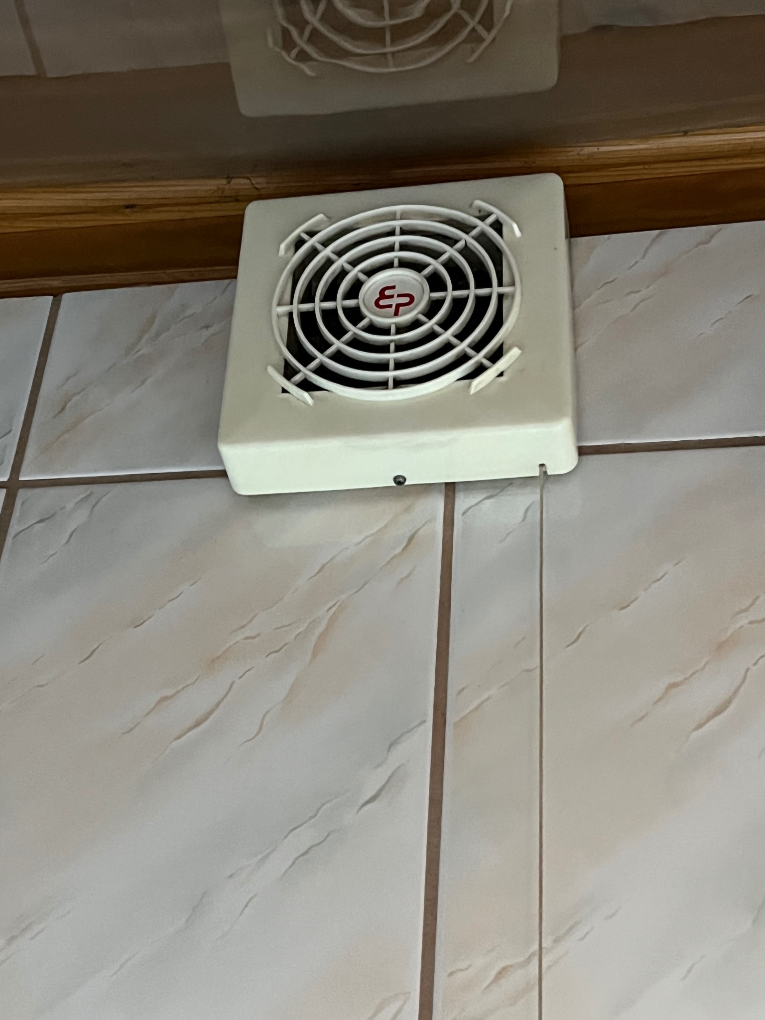 Biały wentylator łazienkowy z czerwonym logo 'EP' na beżowych płytkach ceramicznych, zamontowany pod drewnianą listwą.