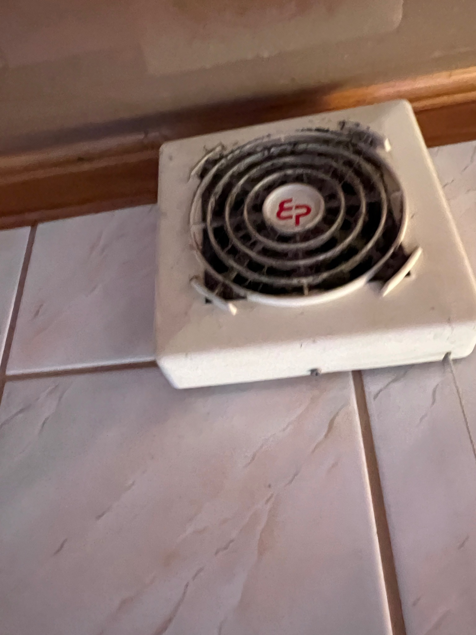 Biały, kwadratowy wentylator łazienkowy z logo 'EP' w czerwonym okręgu, widoczny na kafelkach ściennych, pokryty kurzem.