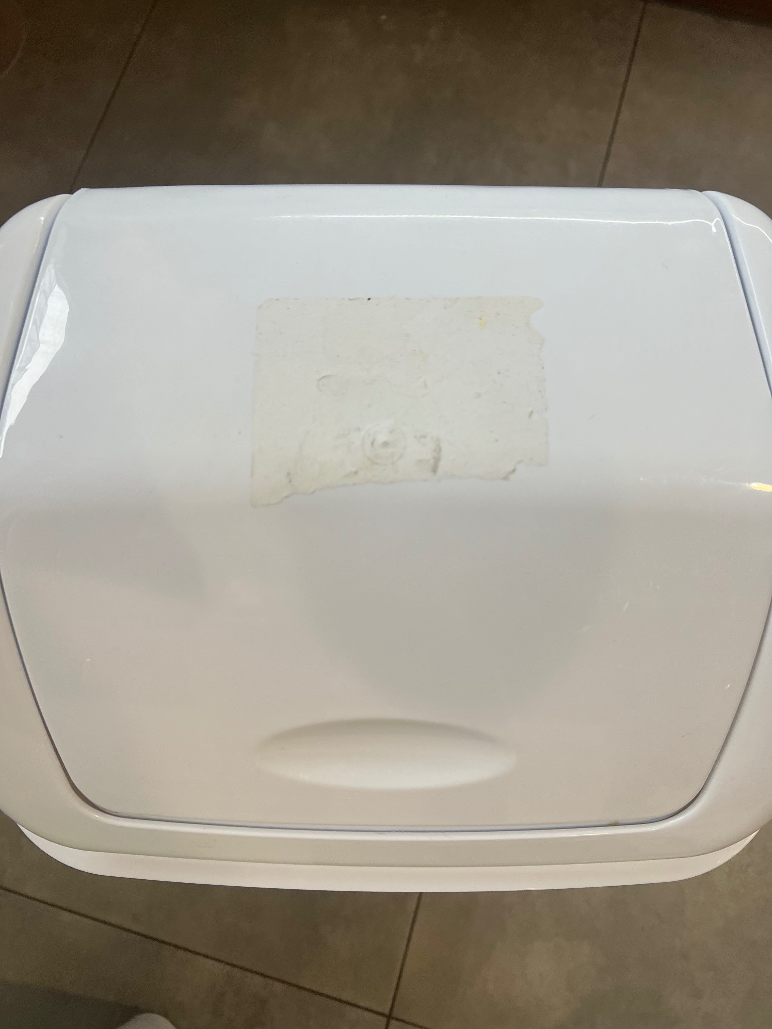 Biały pojemnik na odpady z resztkami kleju po naklejce na pokrywie, widok z góry na brązowe płytki podłogowe.