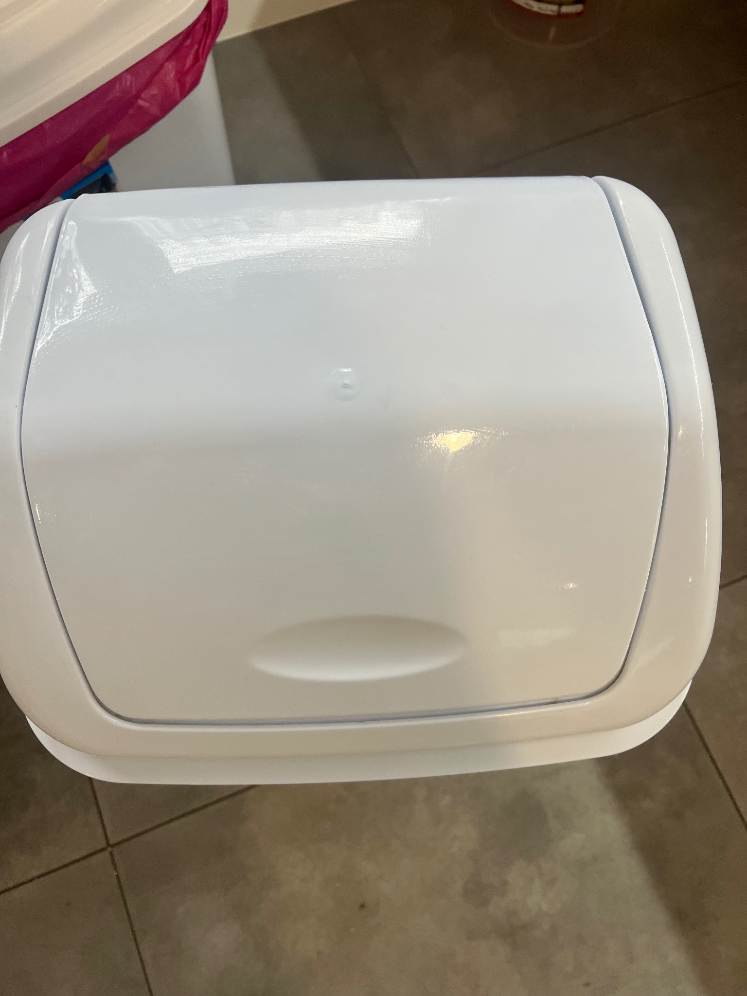 Zamknięty, biały pojemnik na pieluchy na brązowych płytkach podłogowych, widziany z góry.