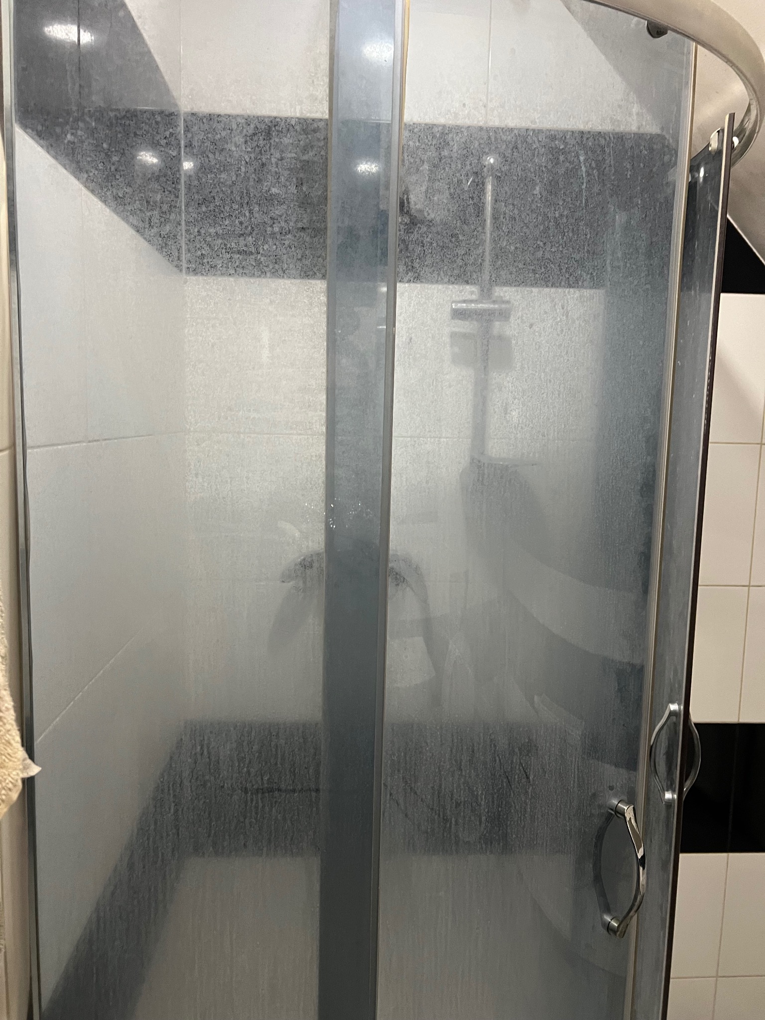 Zabrudzona kabina prysznicowa z widocznymi zaciekami i osadami na szklanych drzwiach i płytkach. Widoczny fragment ręcznika po lewej stronie.