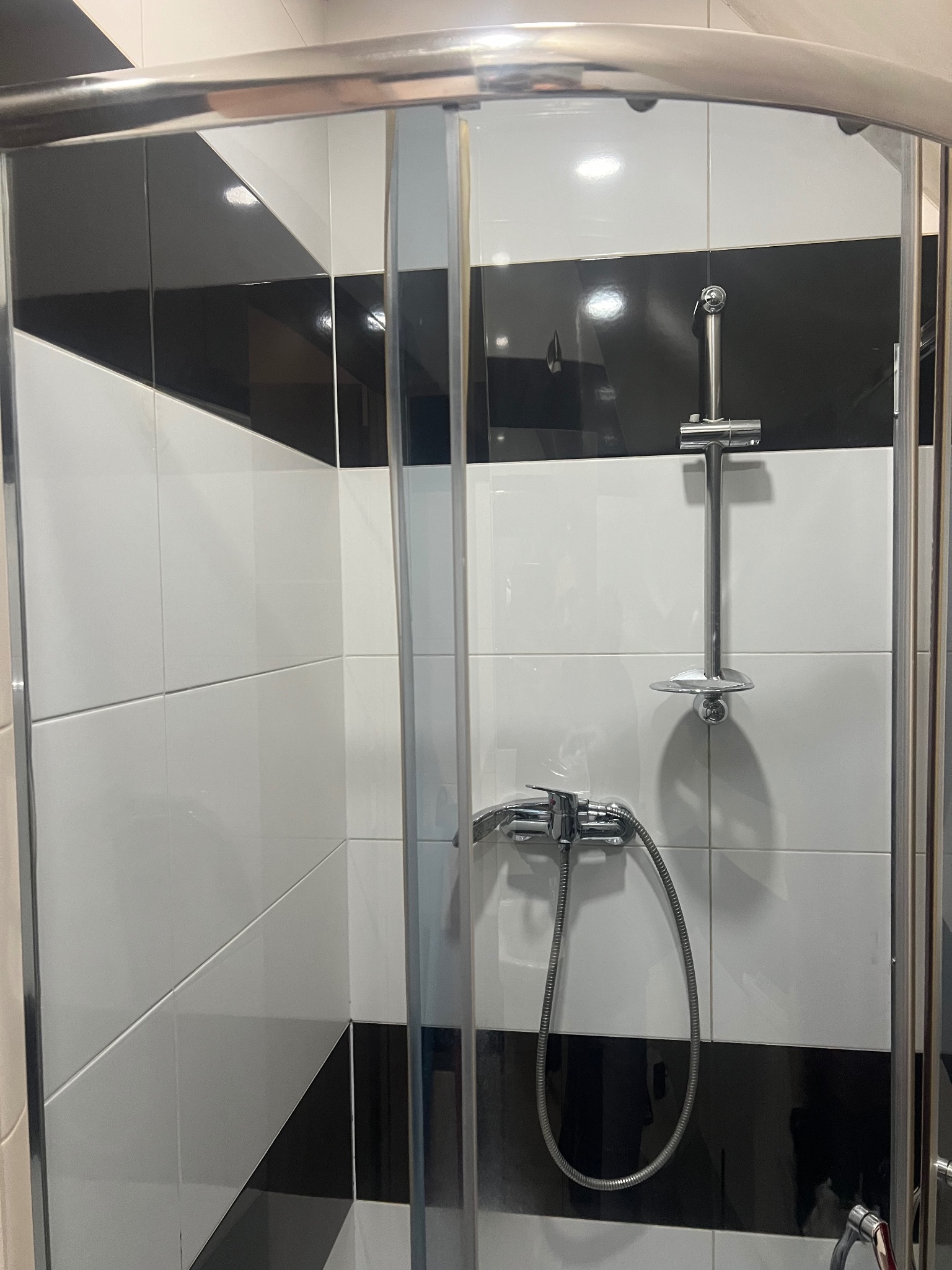 Wnętrze kabiny prysznicowej z chromowaną armaturą i czarno-białymi, geometrycznymi płytkami na ścianach.