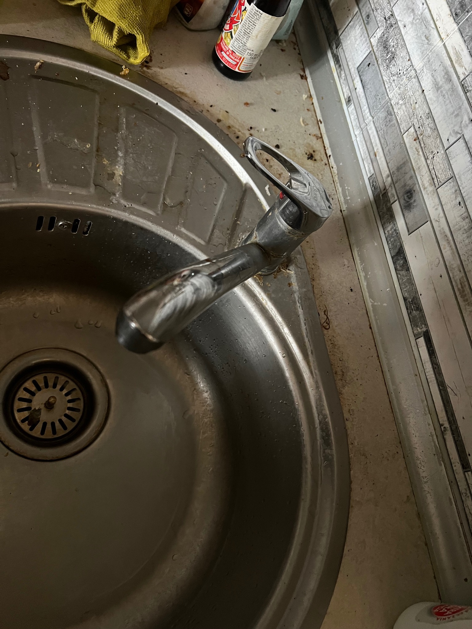 Zanieczyszczony zlew kuchenny ze stali nierdzewnej, widoczne resztki jedzenia i brud na krawędziach zlewu i blacie, obok butelka z sosem i ścierka.