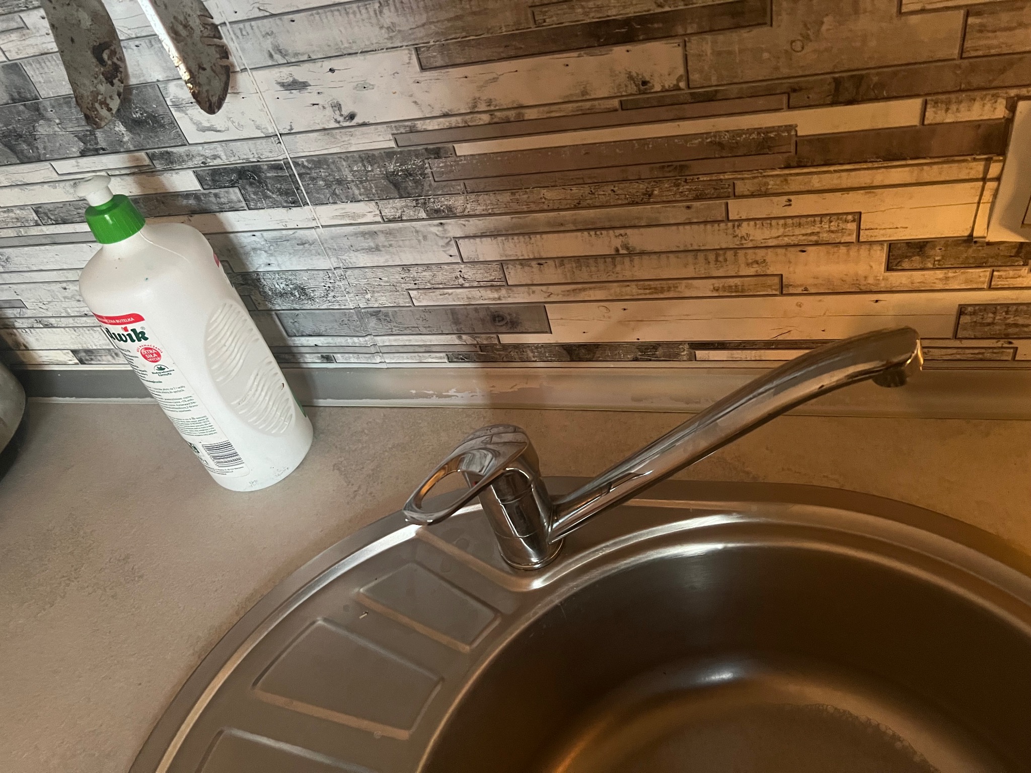 Biała butelka płynu do mycia naczyń stoi obok zlewu ze stali nierdzewnej, na tle ściany wyłożonej panelami imitującymi drewno.