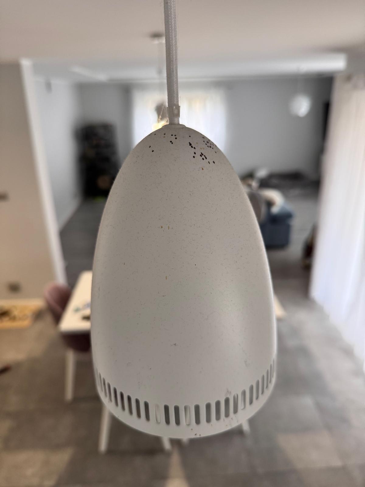 Biała lampa wisząca w kształcie jaja z perforowaną górą i dolną krawędzią, widoczna w tle jadalnia z białym stołem i krzesłami oraz salon z szarą sofą.