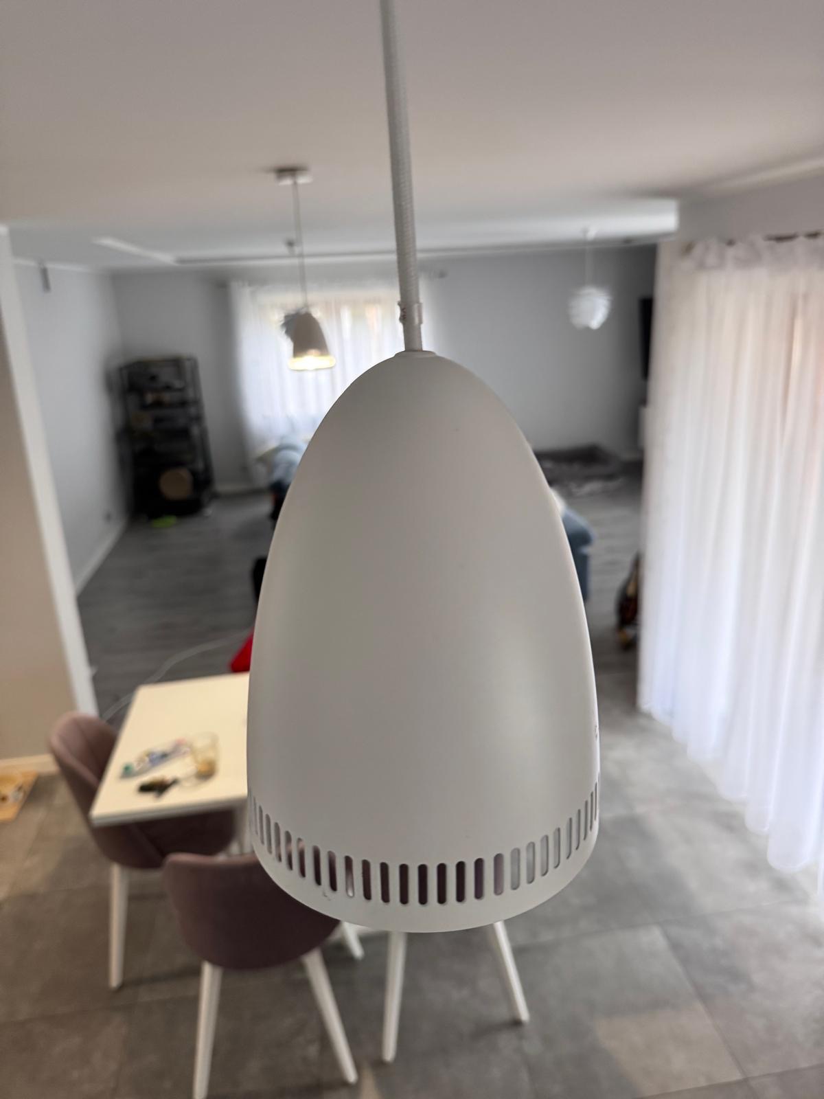 Biała lampa wisząca w kształcie jajka z perforacją u dołu, widok z bliska na tle jadalni z białym stołem i różowymi krzesłami, w tle okno z białymi zasłonami.