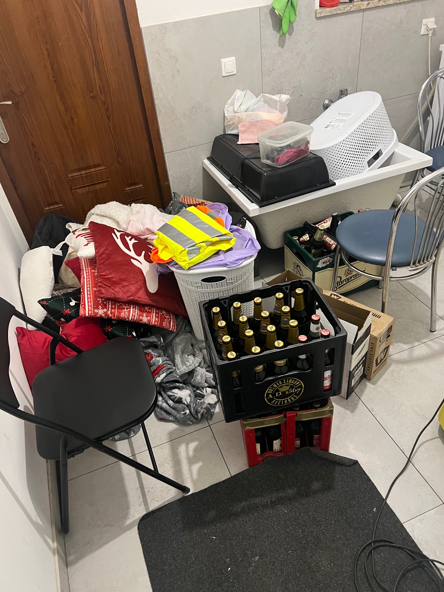 Zagracona łazienka z stertą ubrań, poduszek świątecznych, koszem na pranie, skrzynkami piwa i innymi przedmiotami, tworząca wrażenie chaosu i nieporządku.