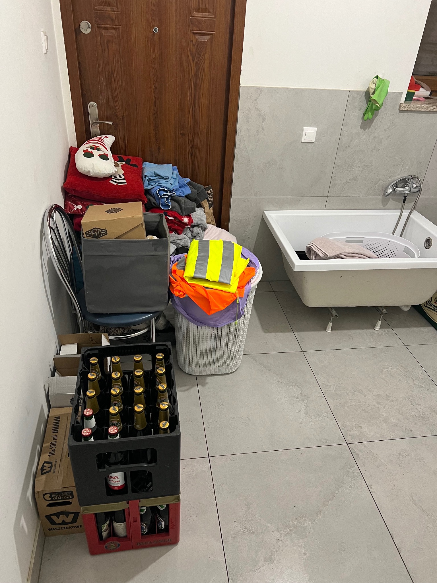 Zagracona łazienka z koszem pełnym ubrań, stertą rzeczy pod ścianą, skrzynką piwa i wanną z praniem.