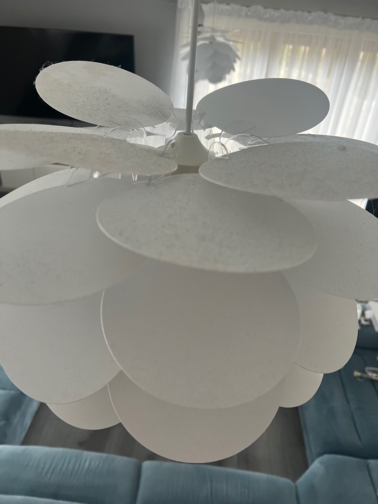 Biała lampa sufitowa z okrągłych, nakładających się na siebie elementów, widoczny kurz i drobne zabrudzenia na niektórych płatkach, na tle zasłon i fragmentu niebieskiej sofy.