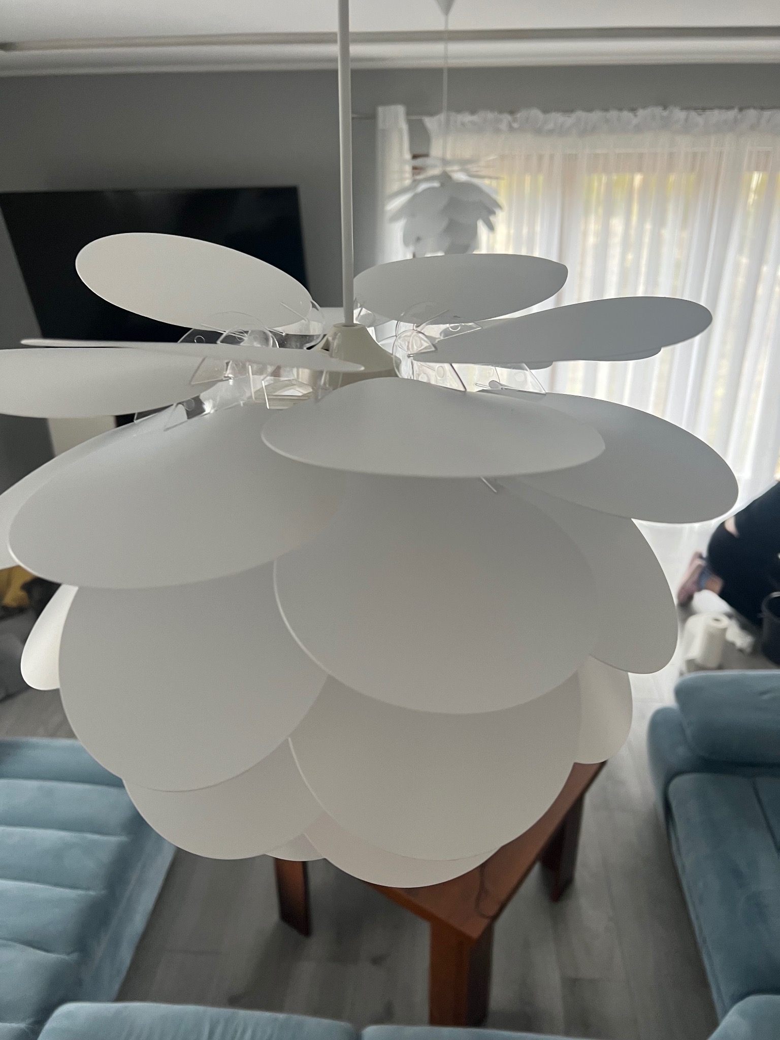 Biała lampa wisząca z kloszami w kształcie płatków, widoczna na tle okna z jasnymi zasłonami i fragmentem niebieskiej sofy.