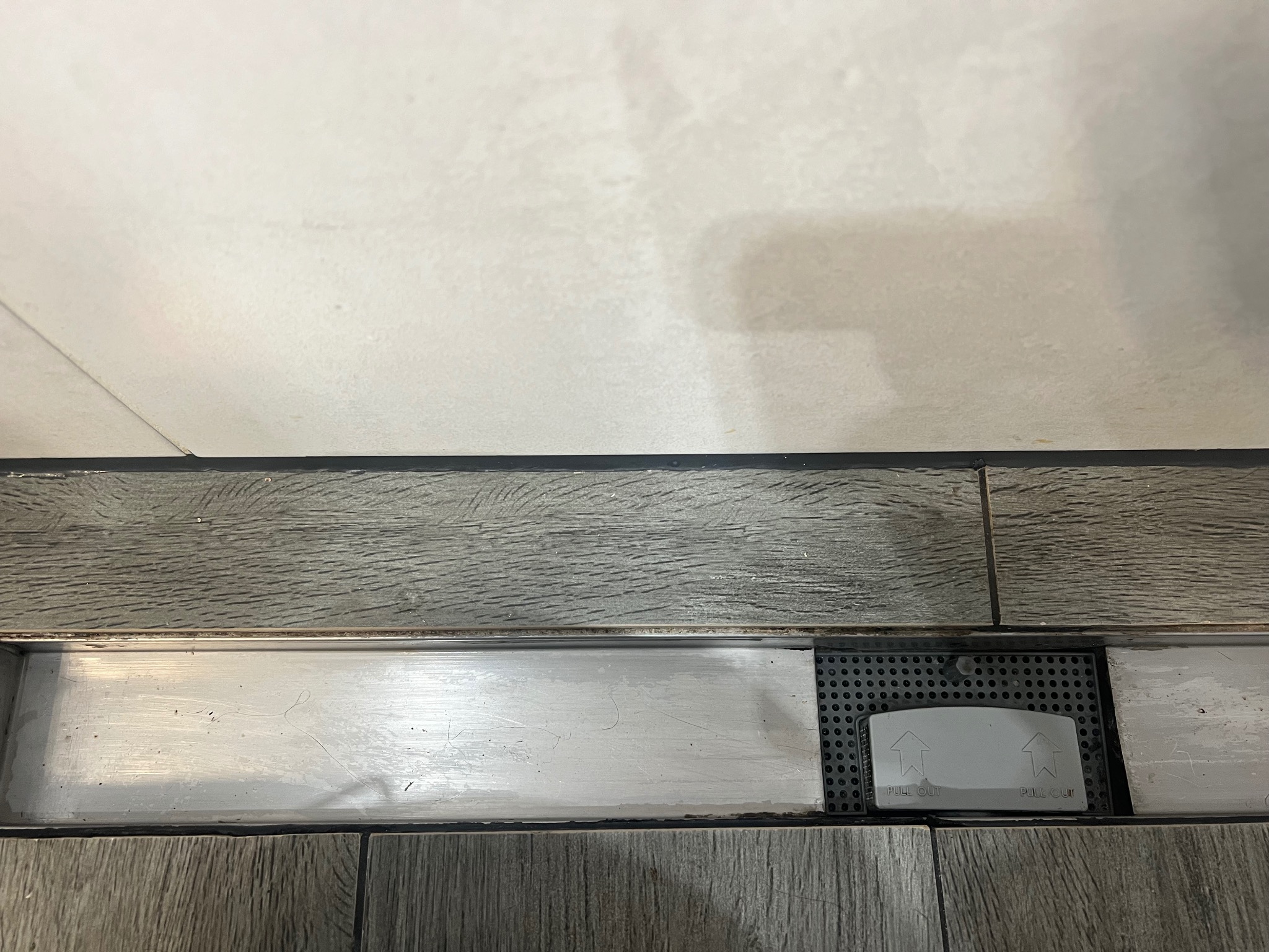 Szczegół odpływu liniowego prysznicowego z szarą kratką i napisem 'PULL OUT', wkomponowanego w płytki imitujące drewno, z widoczną górną częścią ściany wyłożonej jasnymi płytkami.