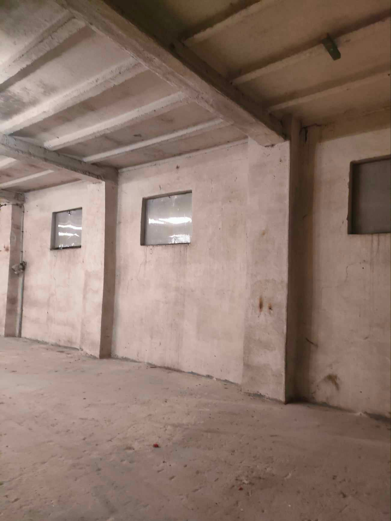 Pusta hala magazynowa z betonowymi ścianami, stropem i trzema oknami z szybami zabezpieczonymi folią.