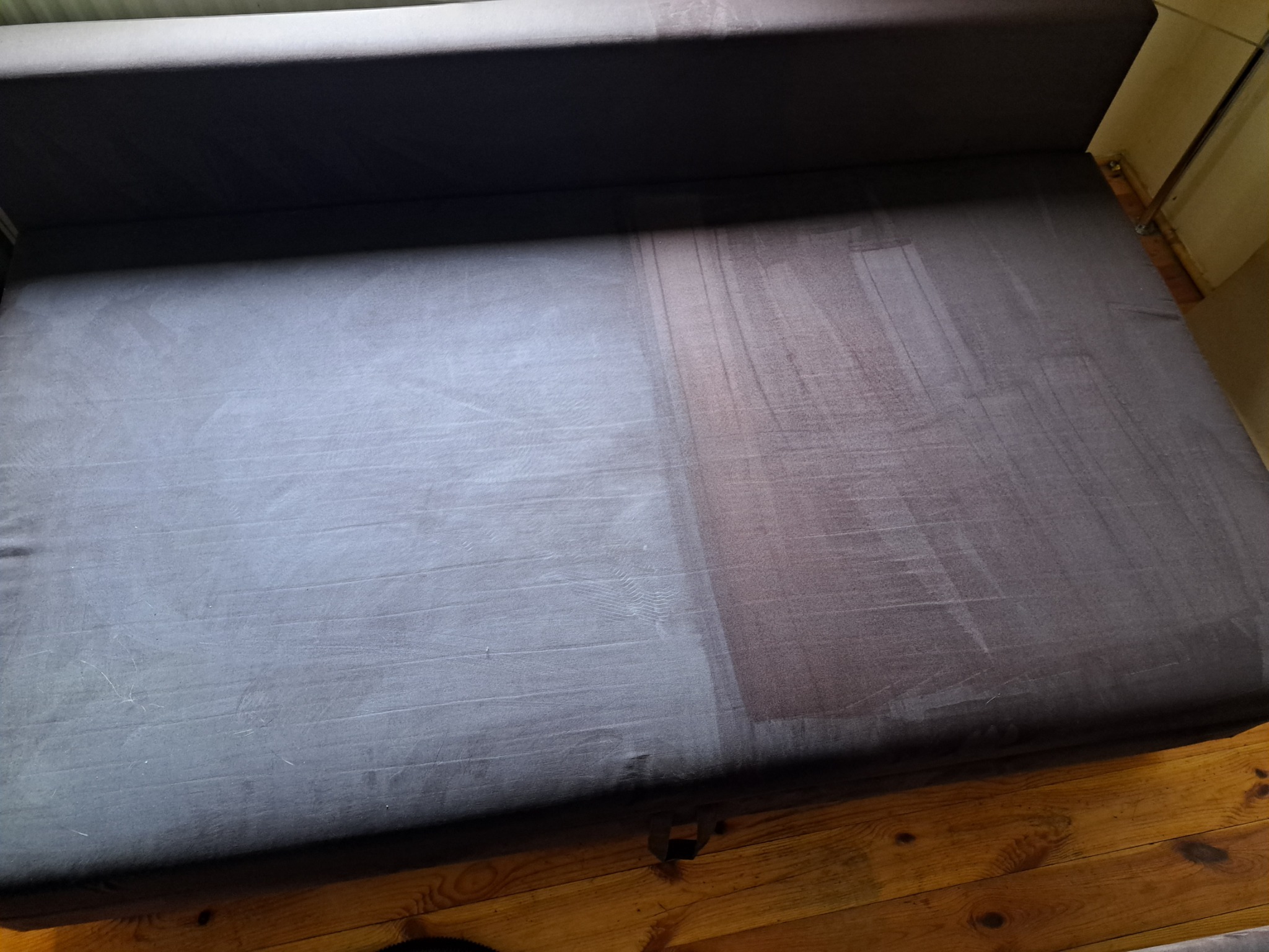 Częściowo wyczyszczona, szara kanapa z widoczną różnicą w kolorze między czystą a brudną powierzchnią, na tle drewnianej podłogi.