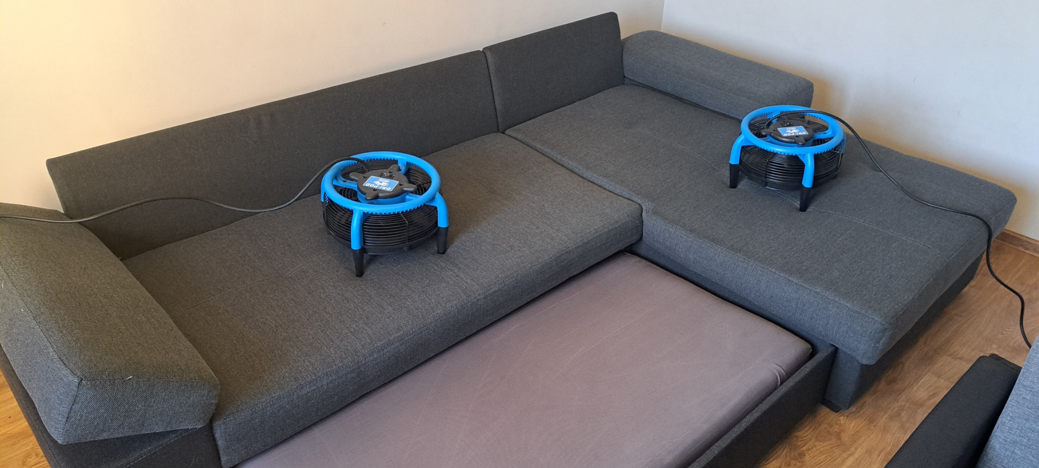 Szara, narożna kanapa z wysuwanym siedziskiem, na której stoją dwa niebiesko-czarne osuszacze powietrza.