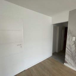 Tomasz Hurkała - Wnętrze po remoncie: białe ściany, drzwi z poziomymi wgłębieniami, panele podłogowe w odcieniach brązu oraz szara ściana z motywem liści palmowych.