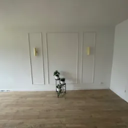 Jasne wnętrze z panelami podłogowymi, białą ścianą z dekoracyjnymi listwami, złotymi kinkietami i stojakiem na kwiaty z trzema doniczkami.