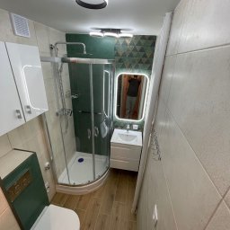 Tomasz Hurkała - Nowoczesna łazienka z kabiną prysznicową narożną, toaletą, umywalką z szafką i podświetlanym lustrem. Na ścianach beżowe płytki, na podłodze panele imitujące drewno, akcenty w kolorze ciemnej zieleni.