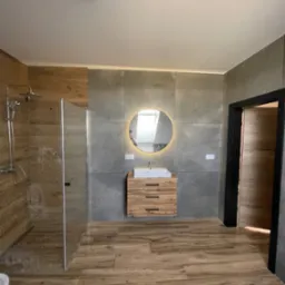 Nowoczesna łazienka z kabiną prysznicową, drewnopodobną podłogą i szarą ścianą z okrągłym lustrem z podświetleniem LED nad drewnianą szafką z umywalką.