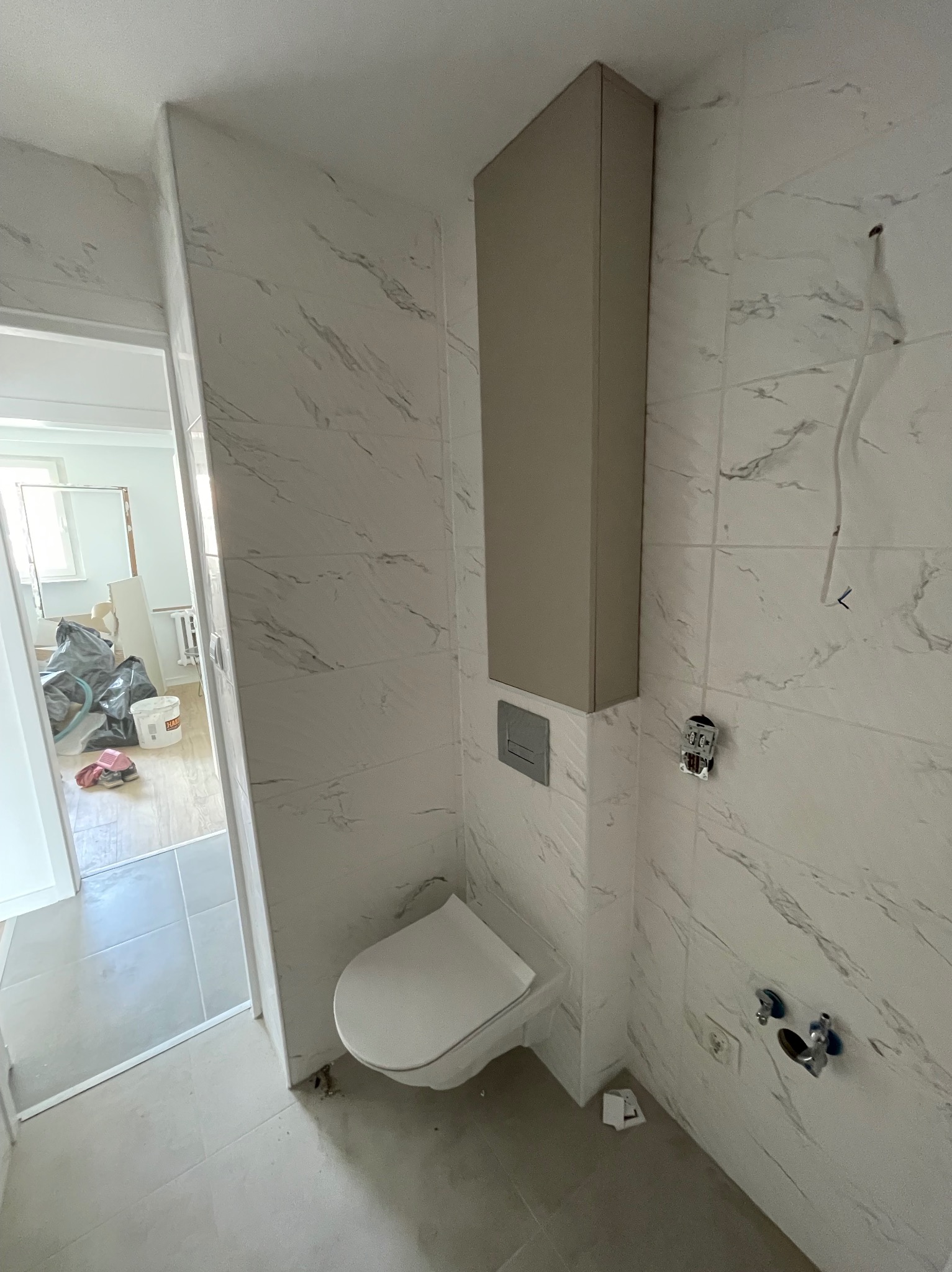 Łazienka w trakcie remontu: białe płytki imitujące marmur na ścianach, wisząca szafka nad toaletą, widoczne niedokończone instalacje elektryczne i hydrauliczne.