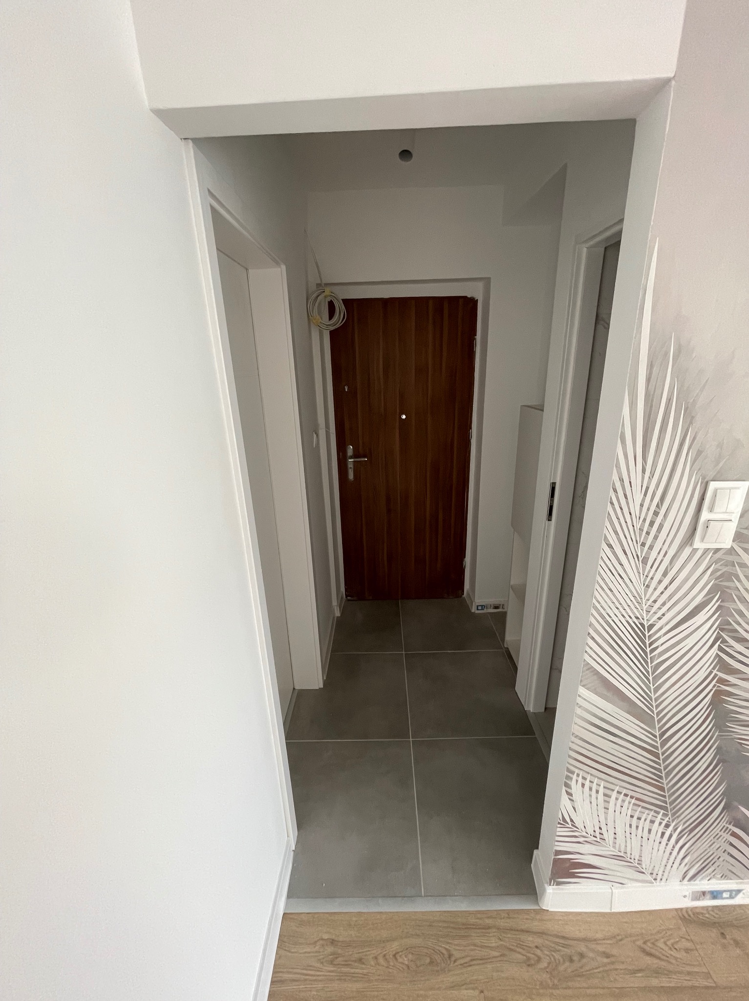 Wnętrze korytarza po remoncie: szare płytki podłogowe, białe ściany, drewniane drzwi wejściowe, tapeta z motywem liści palmowych na fragmencie ściany, widoczne kable wystające ze ściany.