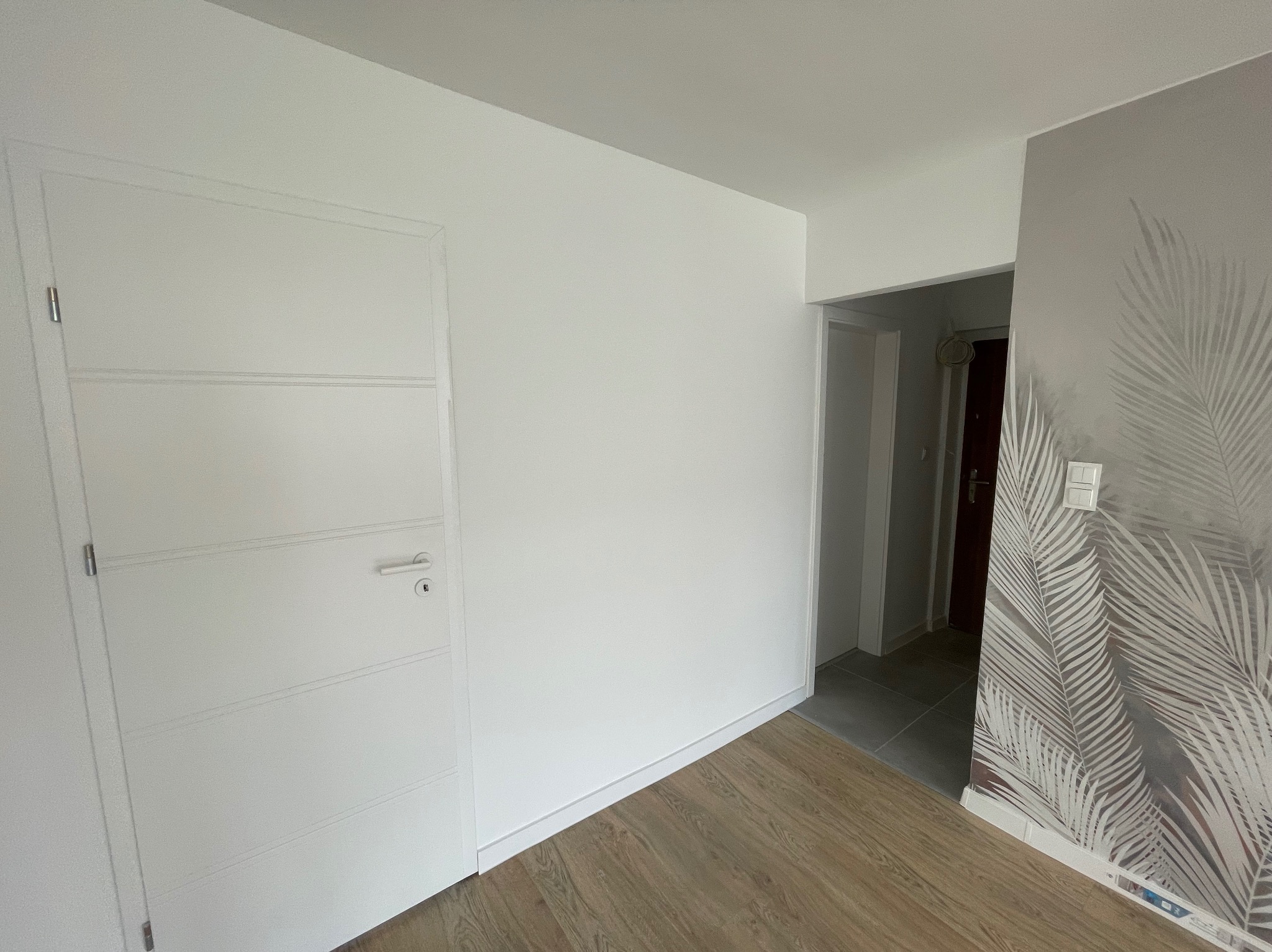 Wnętrze po remoncie: białe ściany, drzwi z poziomymi wgłębieniami, panele podłogowe w odcieniach brązu oraz szara ściana z motywem liści palmowych.