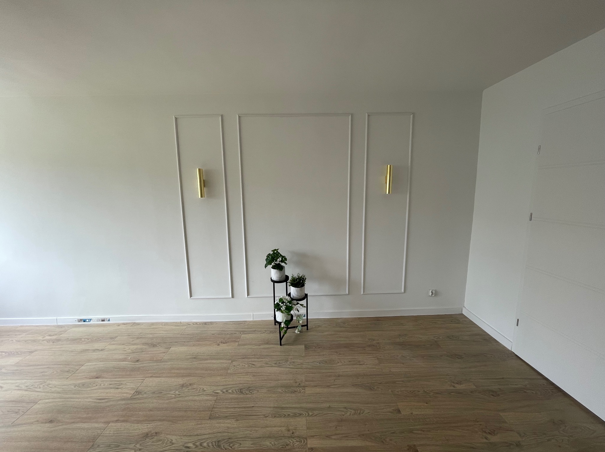 Jasne wnętrze z panelami podłogowymi, białą ścianą z dekoracyjnymi listwami, złotymi kinkietami i stojakiem na kwiaty z trzema doniczkami.