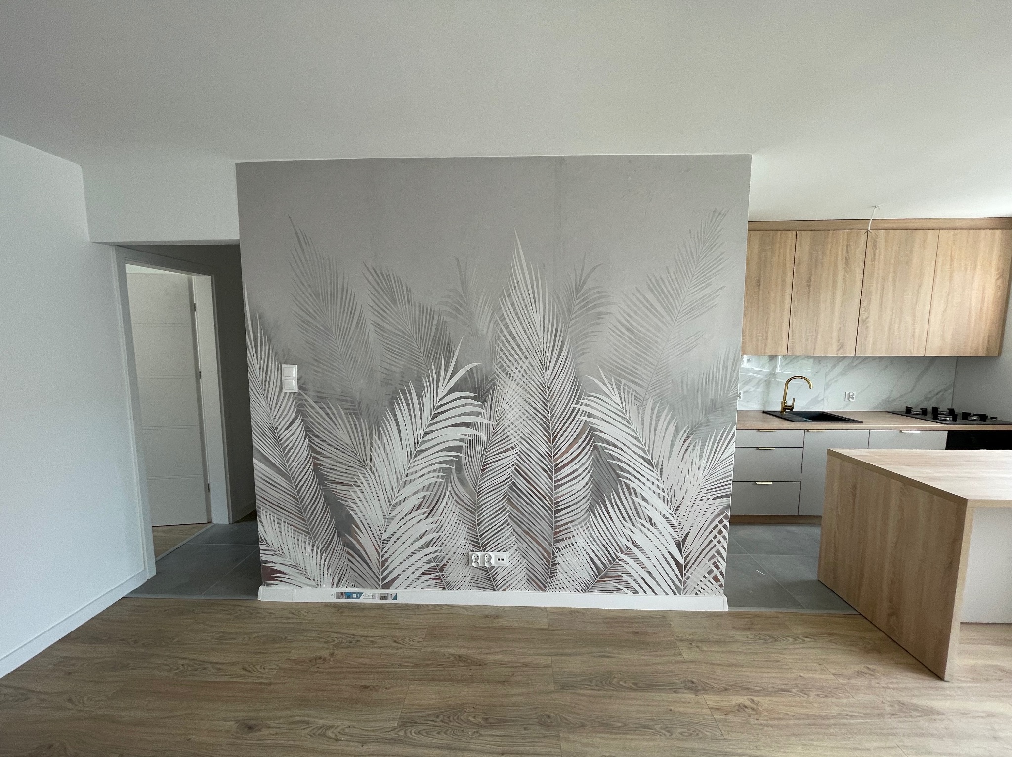 Ściana w mieszkaniu wykończona tapetą z motywem liści palmowych, widoczny fragment kuchni z drewnianymi szafkami i blatem, podłoga z paneli imitujących drewno.