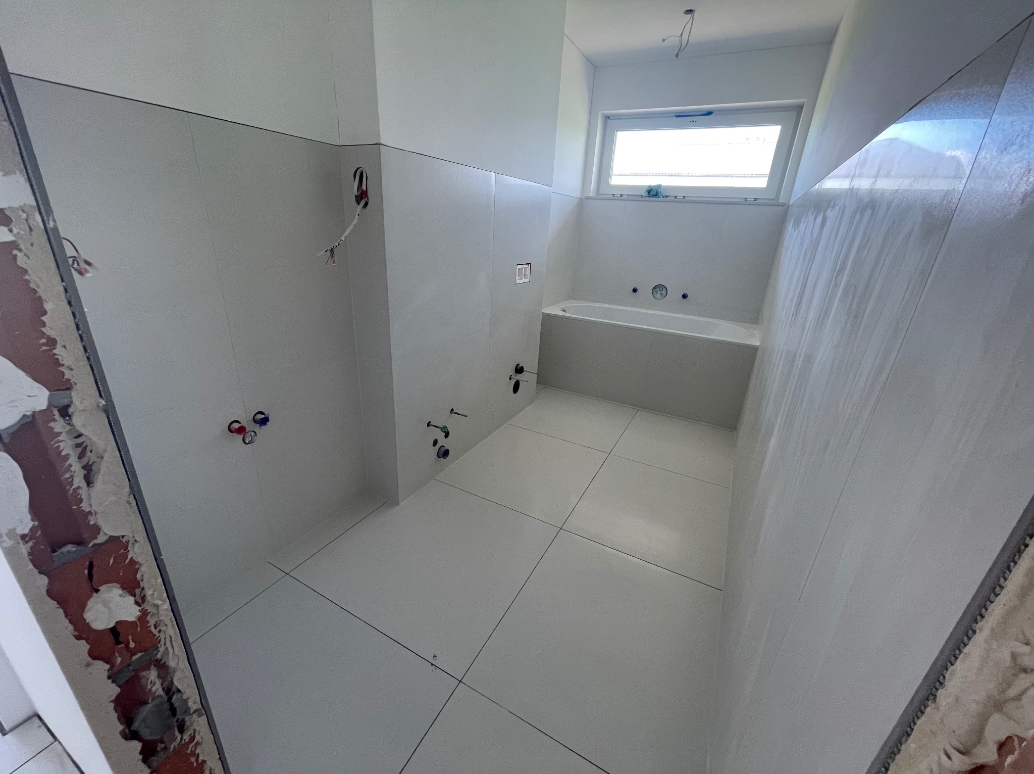 Wnętrze łazienki w trakcie remontu z ułożonymi białymi płytkami na podłodze i ścianach, widoczne wystające instalacje wodne i elektryczne, oraz fragment ściany z cegły przed wykończeniem.