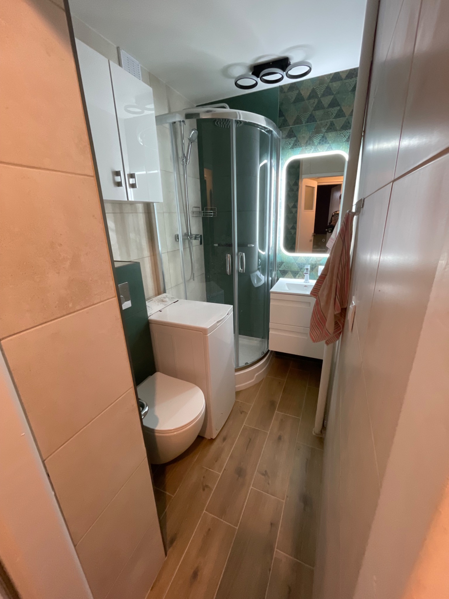 Nowoczesna, wąska łazienka z kabiną prysznicową z ciemną ramą, białą umywalką z podświetlanym lustrem, toaletą typu kompakt i pralką, wykończona płytkami imitującymi drewno i zieloną ścianą...