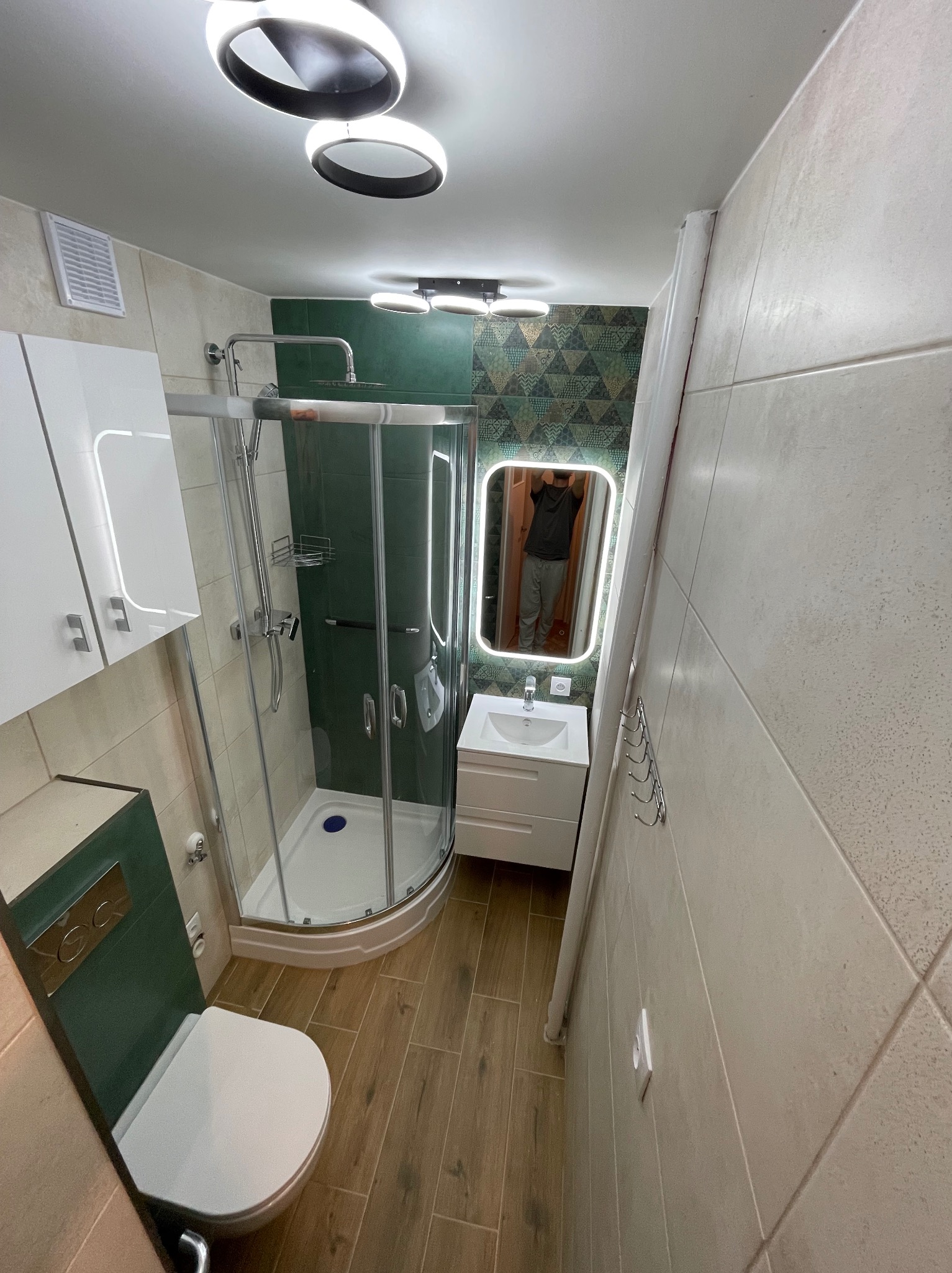 Nowoczesna łazienka z kabiną prysznicową narożną, toaletą, umywalką z szafką i podświetlanym lustrem. Na ścianach beżowe płytki, na podłodze panele imitujące drewno, akcenty w kolorze ciemnej zieleni.