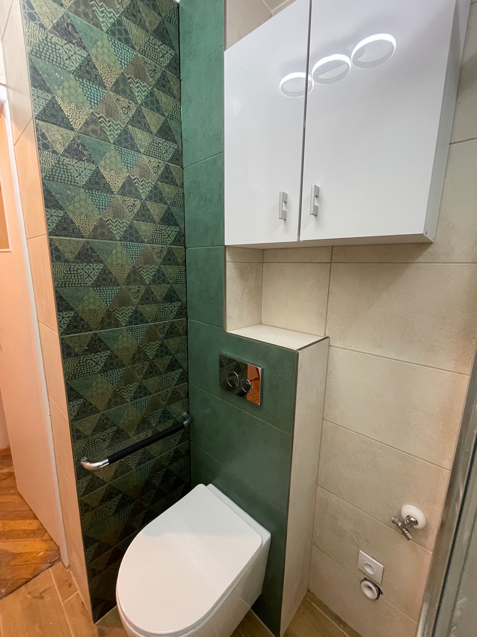 Wykończona toaleta z wzorzystymi, zielono-brązowymi płytkami na ścianie, białą wiszącą miską WC z chromowanym uchwytem dla niepełnosprawnych i szafką wiszącą nad nią, kremowe płytki na pozostałych...