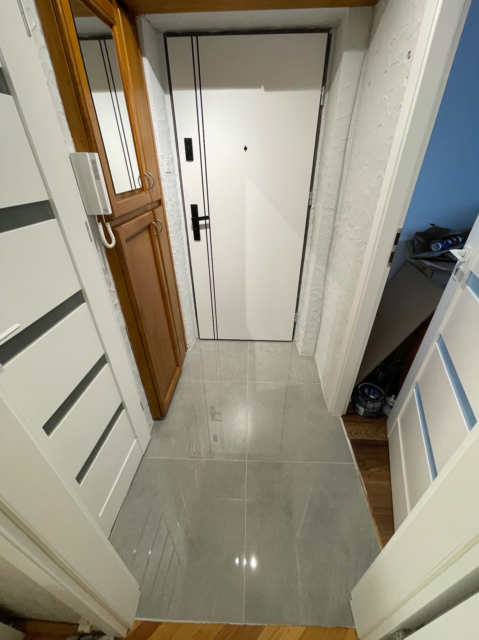 Korytarz po remoncie z błyszczącą, szarą podłogą z płytek, białe drzwi wejściowe z czarnymi akcentami i widoczne drzwi do spiżarni z resztkami materiałów budowlanych.