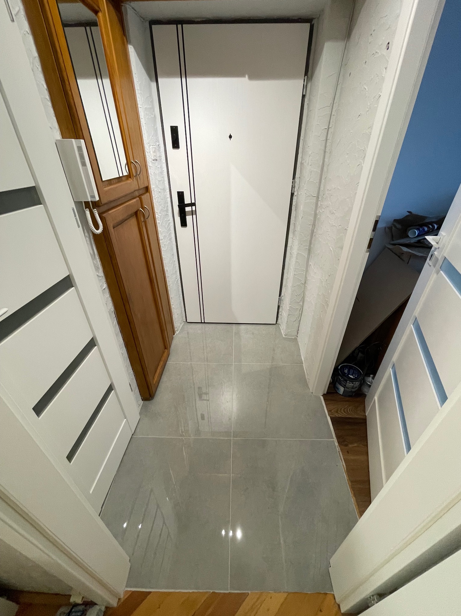 Wykończony korytarz z szarą, błyszczącą terakotą, białe drzwi wejściowe z czarnymi paskami, drewniana szafa z lustrem i biały domofon, widok z wnętrza mieszkania.