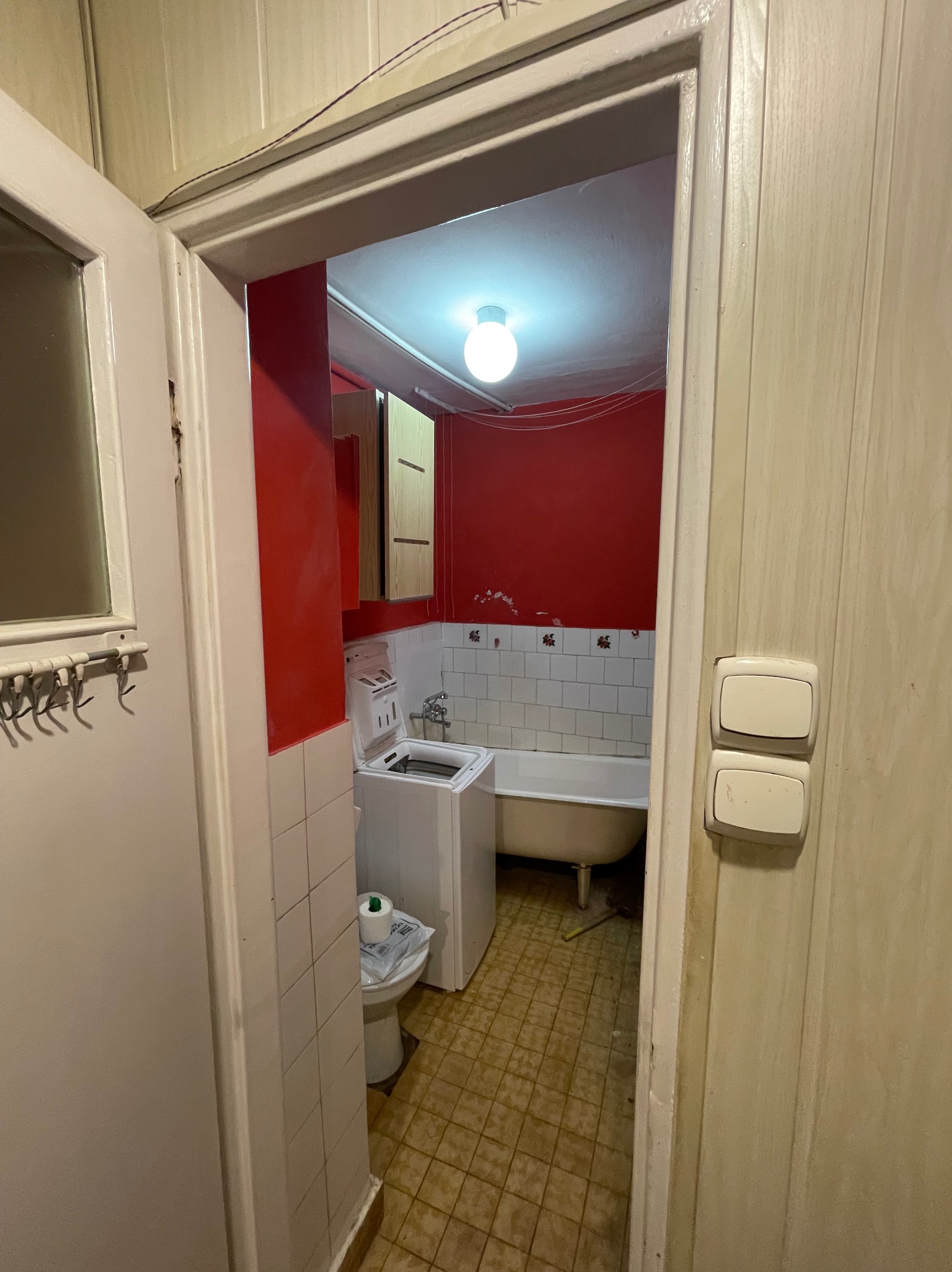 Wnętrze łazienki z czerwonymi ścianami, białą wanną na nóżkach i pralką, widok z korytarza przez otwarte drzwi, z widocznymi starymi płytkami i osprzętem.