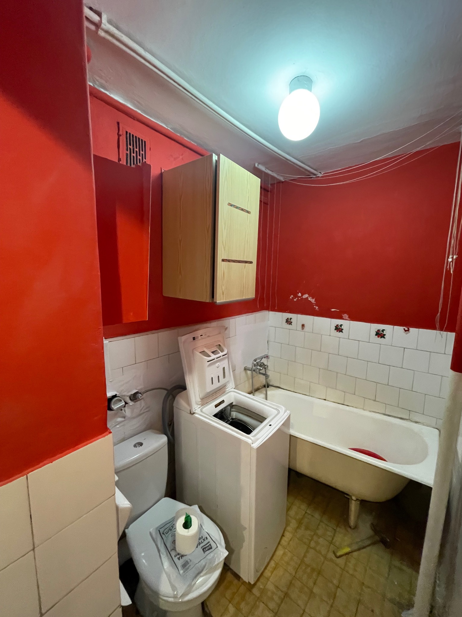 Mała łazienka z czerwonymi ścianami, białą wanną na nóżkach, pralką z otwartą klapą, toaletą z rolką papieru, szafką wiszącą i kafelkami z motywem kwiatowym.