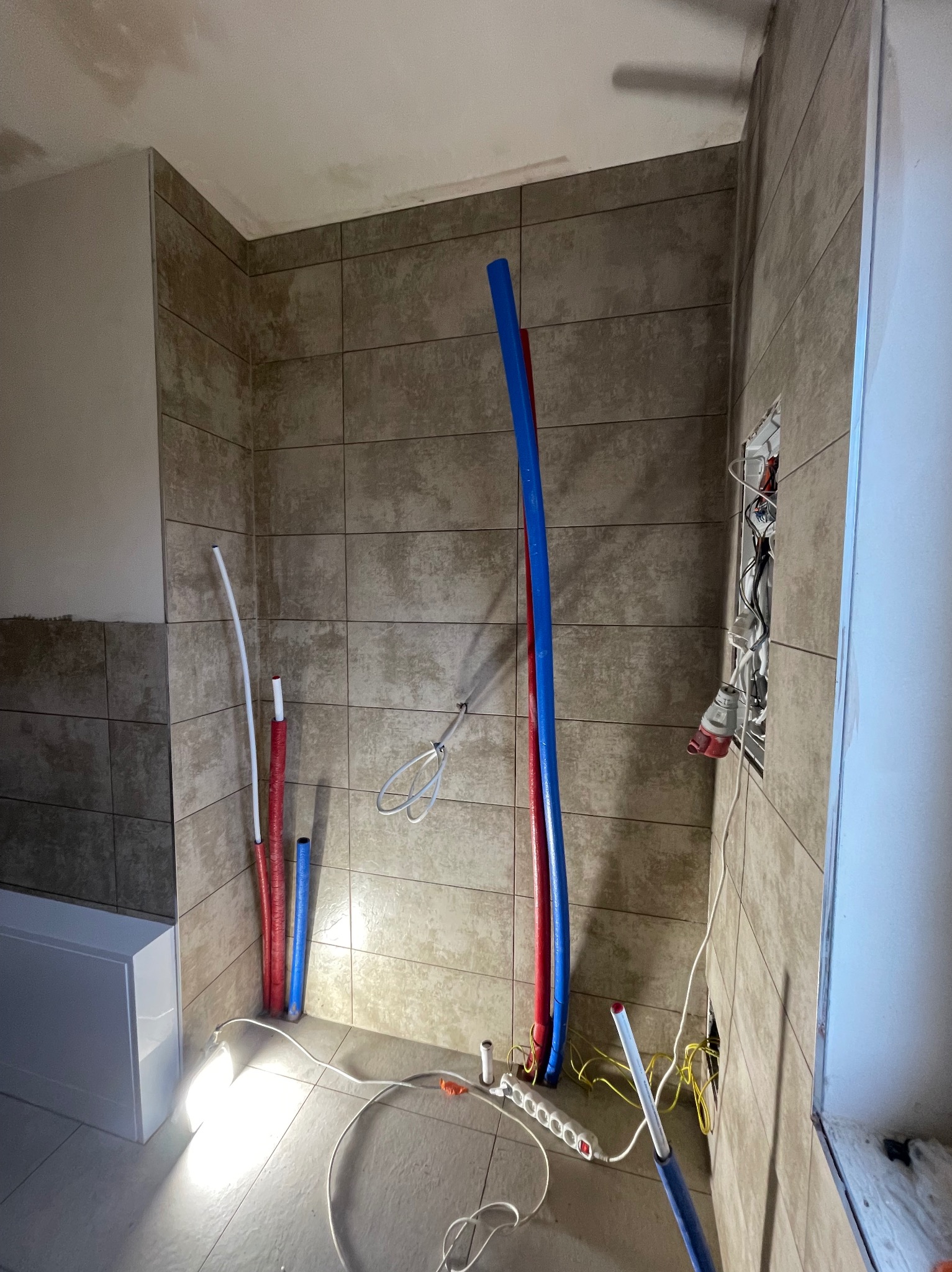 Łazienka w trakcie remontu: ułożone płytki imitujące kamień, wystające rury instalacji wodnej w kolorach niebieskim i czerwonym, widoczne przewody elektryczne i przedłużacz. Oświetlenie robocze...