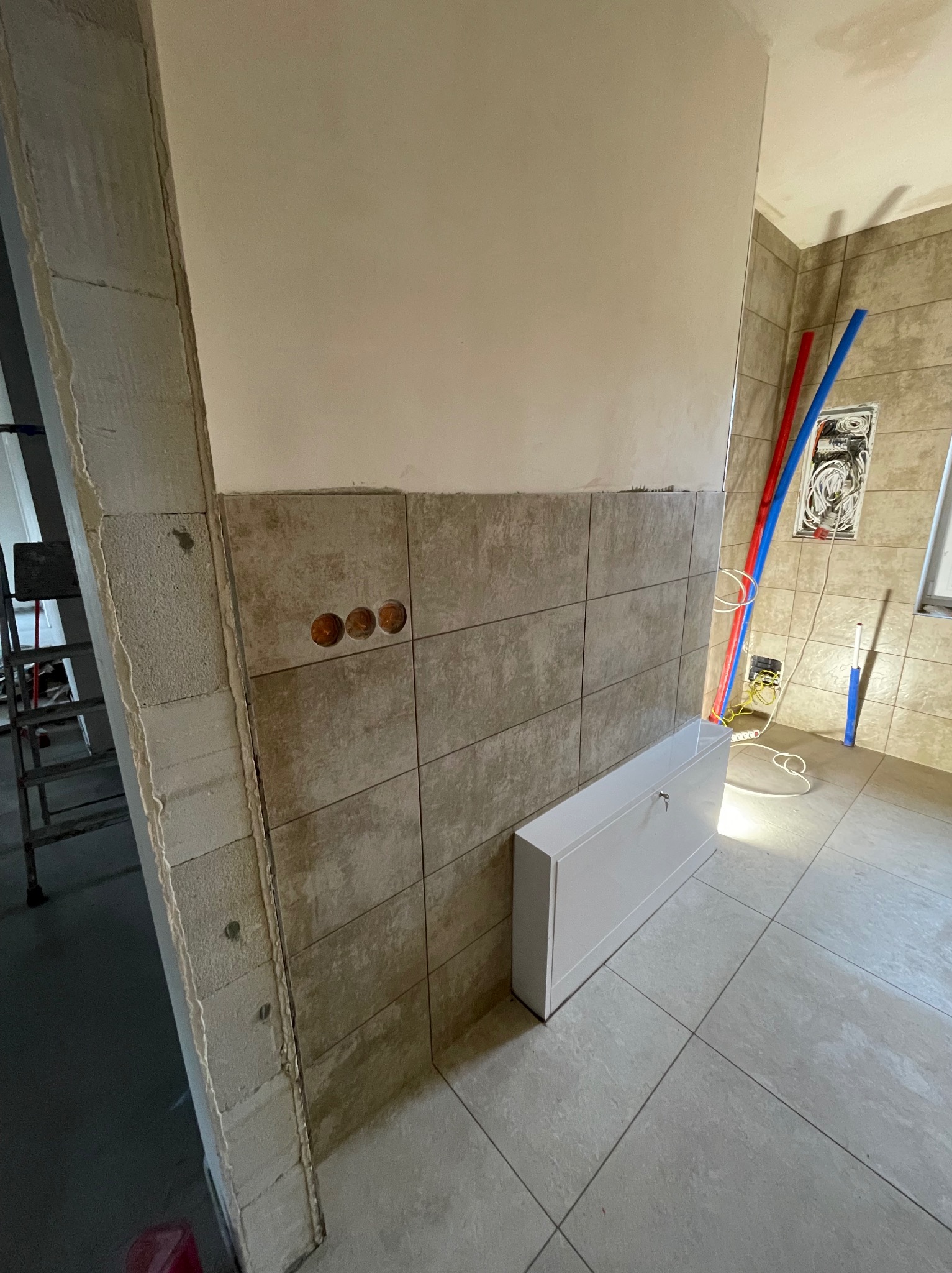 Ściana łazienki w trakcie remontu: częściowo ułożone płytki ceramiczne w odcieniach beżu, widoczne puszki elektryczne bez osprzętu, fragment ściany z surowego betonu komórkowego, biała szafka...