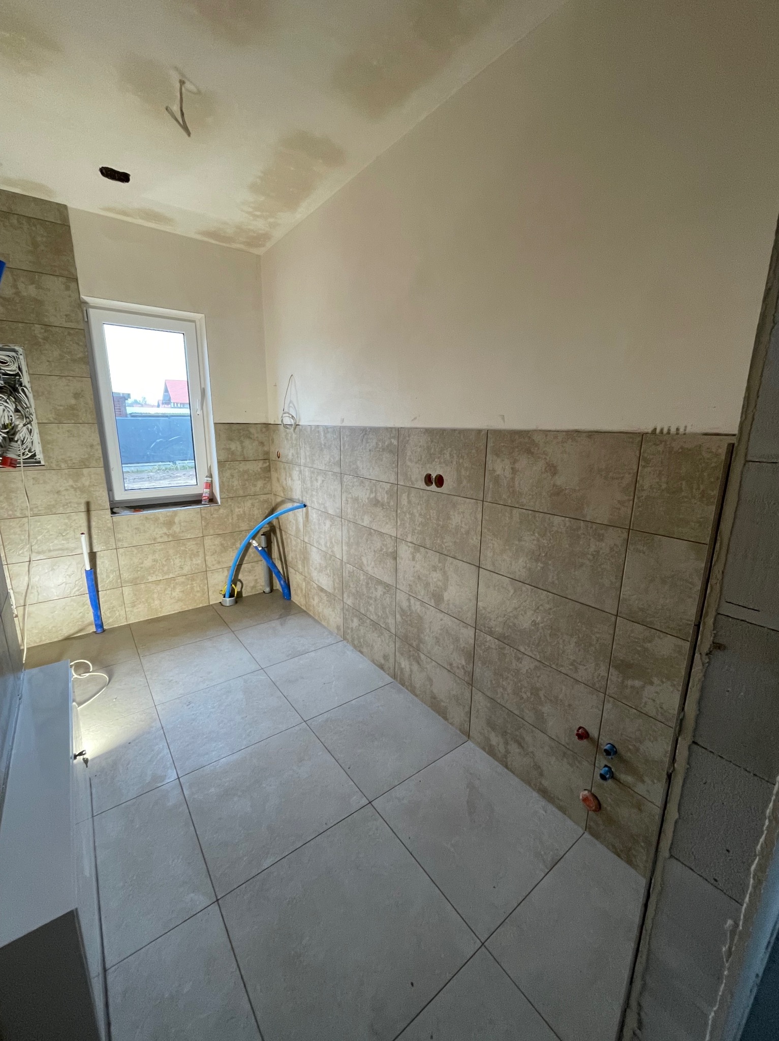 Świeżo wykafelkowana łazienka z beżowymi płytkami na ścianach i podłodze, widoczne rury doprowadzające wodę, sufit wymaga jeszcze wykończenia.
