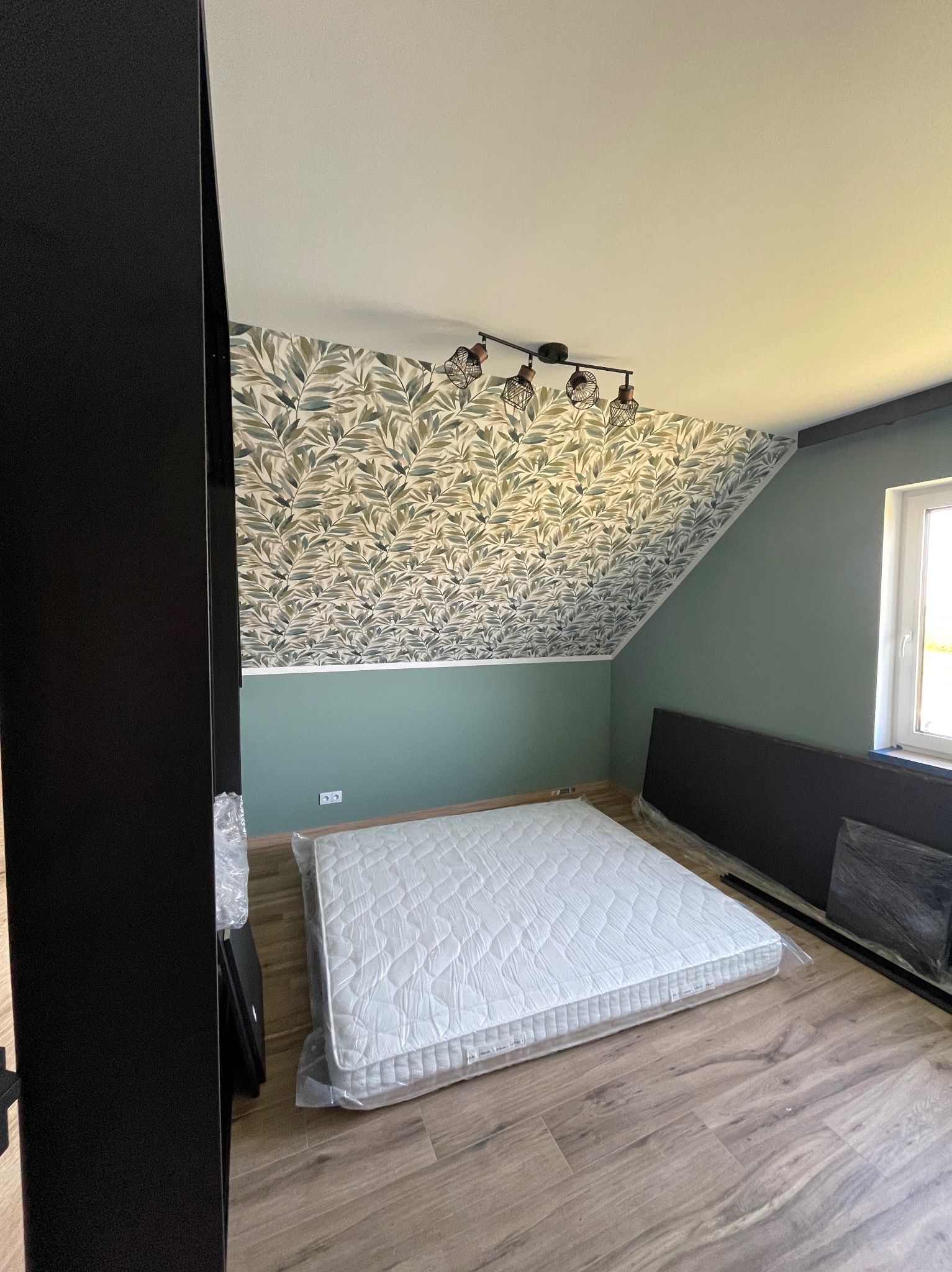 Pokój na poddaszu po remoncie: wzorzysta tapeta na skosach, zielone ściany, panele podłogowe. Nowy materac w folii czeka na rozpakowanie. Nowoczesne oświetlenie.