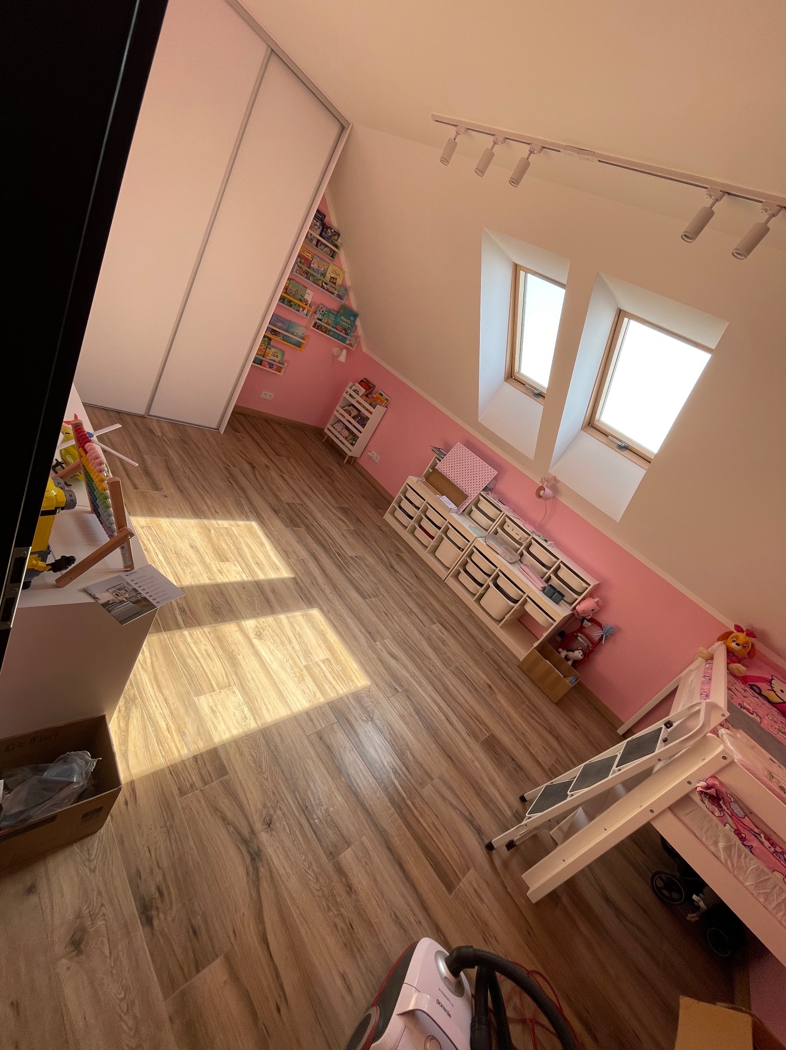 Jasne panele podłogowe w pokoju dziecięcym na poddaszu w Oławie. Widoczne okna dachowe, różowe ściany i białe meble. Uwagę zwraca drewniana podłoga.