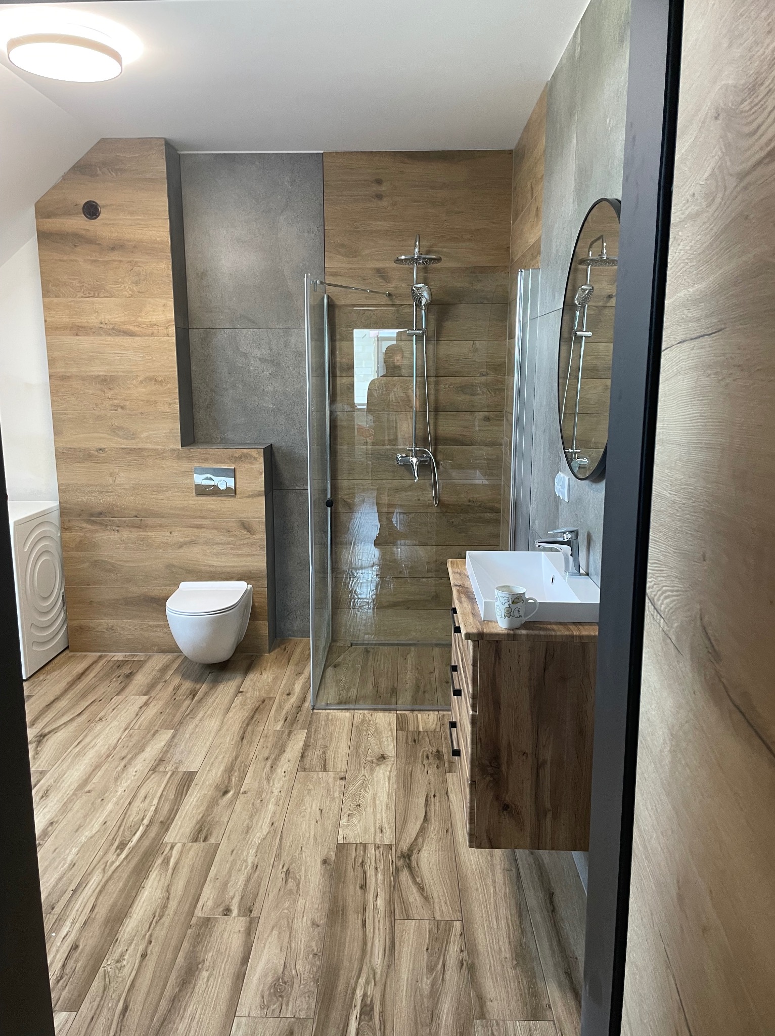 Nowoczesna łazienka z kabiną prysznicową typu walk-in, drewnopodobnymi płytkami na podłodze i ścianach oraz szarą strefą. Umywalka z szafką i okrągłe lustro.