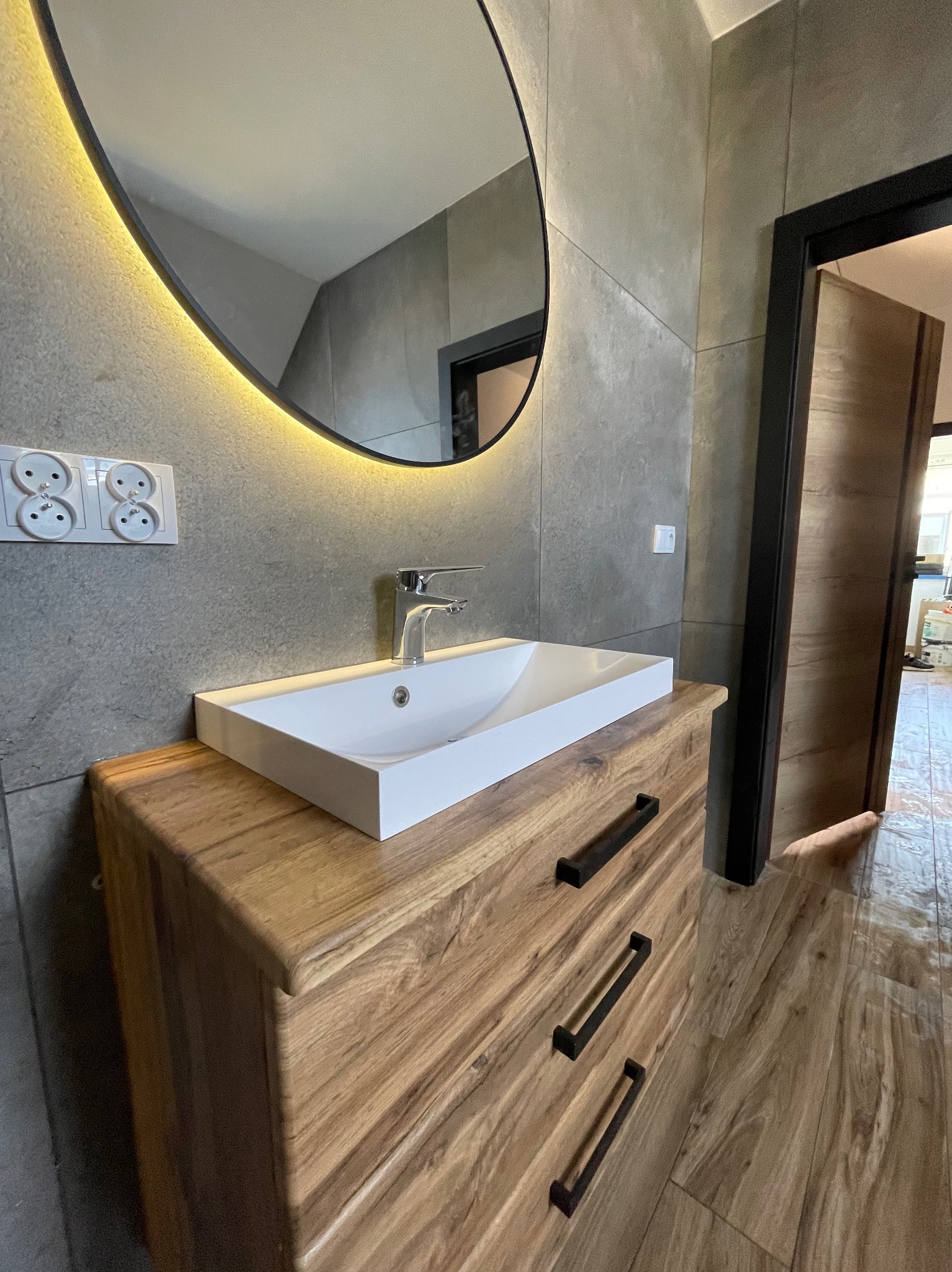 Nowoczesna łazienka z umywalką nablatową na drewnianej szafce, owalne lustro z podświetleniem LED i szare płytki imitujące kamień na ścianach.