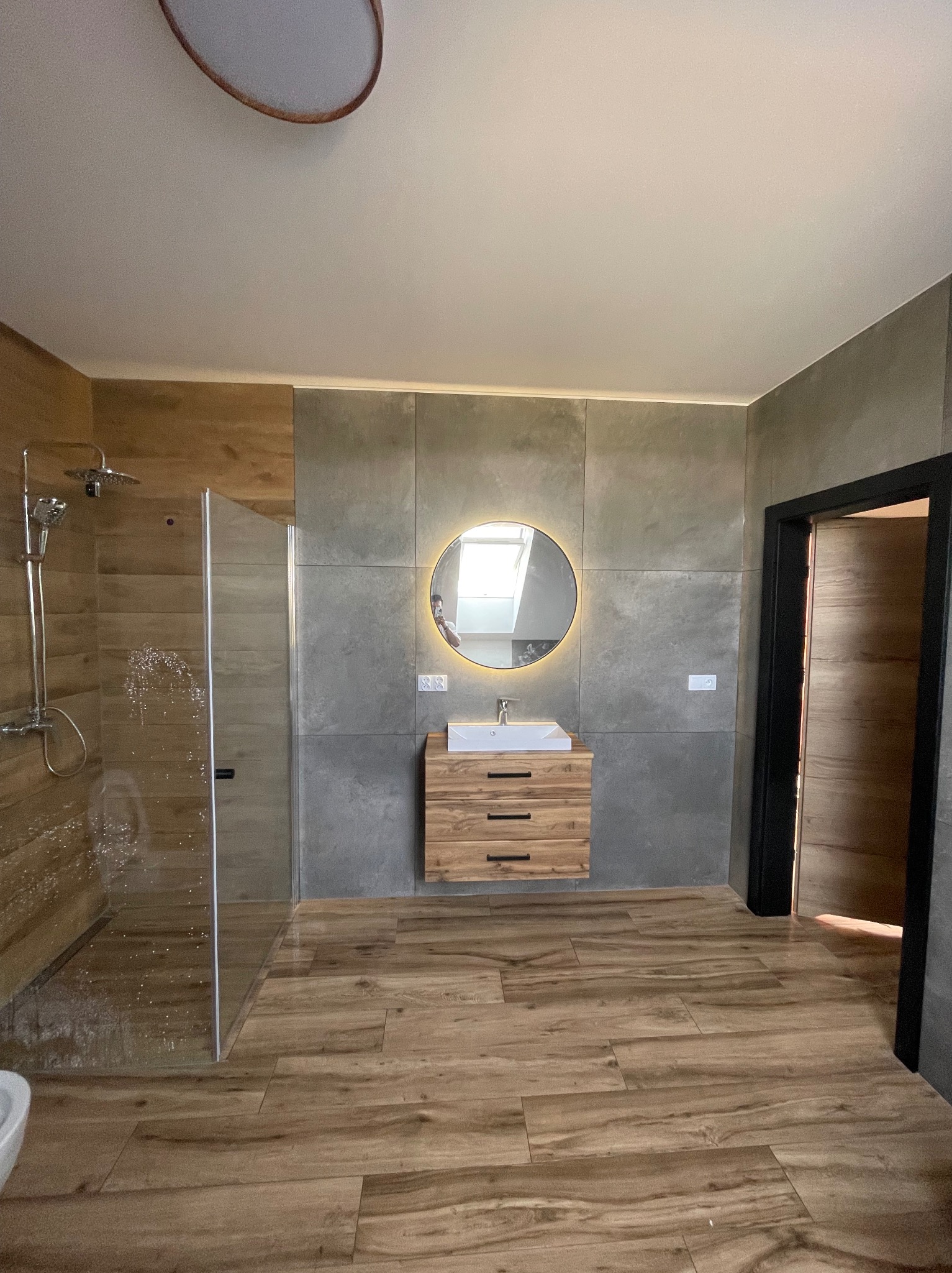 Nowoczesna łazienka z kabiną prysznicową, drewnopodobną podłogą i szarą ścianą z okrągłym lustrem z podświetleniem LED nad drewnianą szafką z umywalką.
