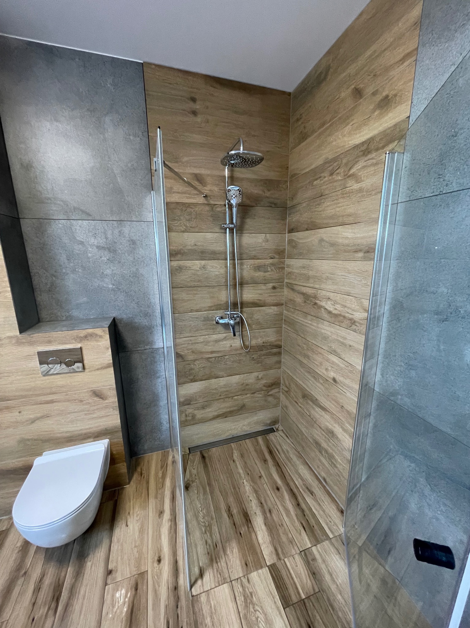 Nowoczesna kabina prysznicowa z deszczownicą, wykończona płytkami imitującymi drewno i szary beton. Uwagę zwraca odpływ liniowy w podłodze.