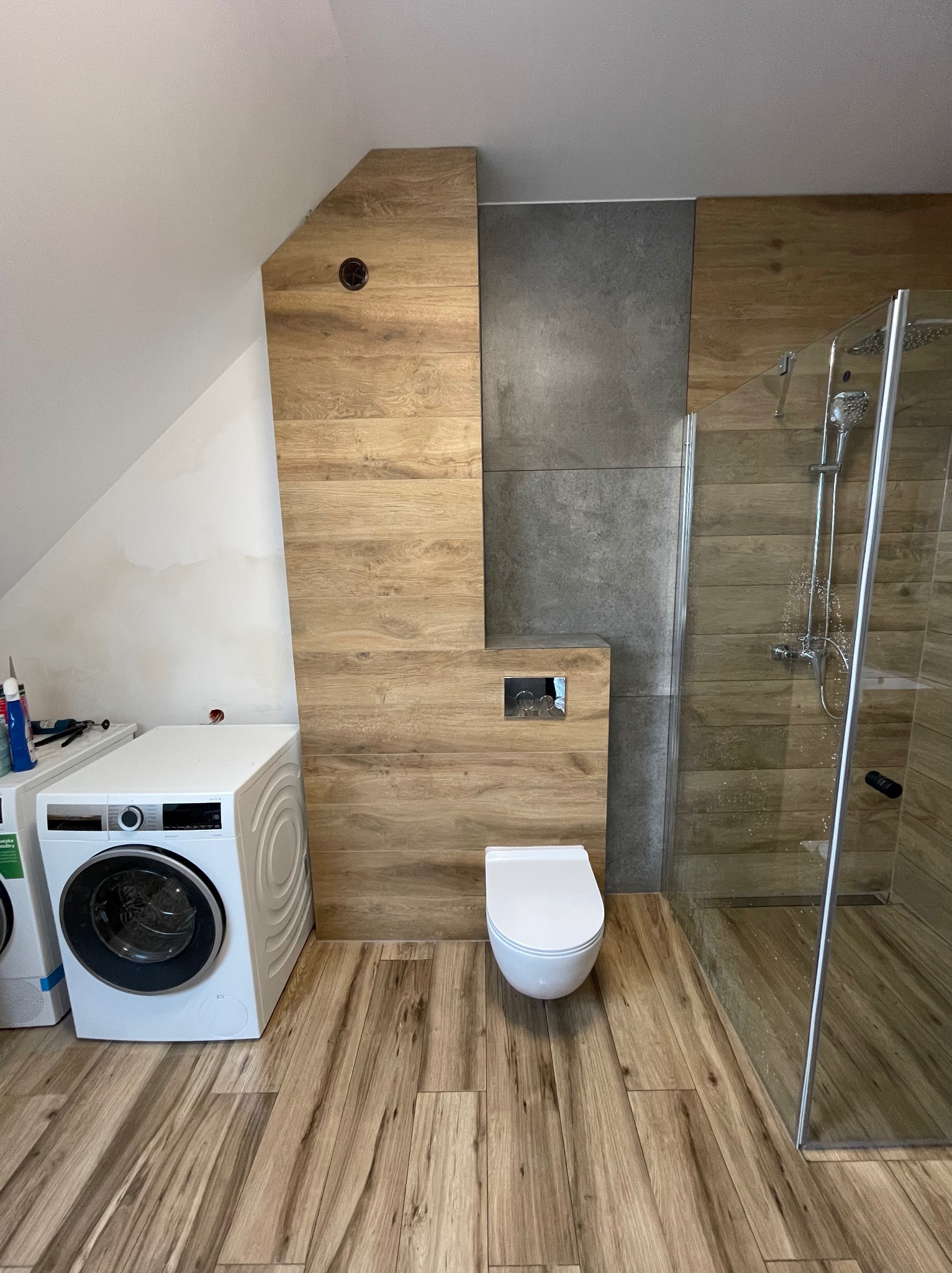 Nowoczesna łazienka na poddaszu z drewnopodobnymi płytkami, kabiną prysznicową i pralką. Uwagę zwraca nietypowe wykończenie ściany z ukrytym systemem spłukiwania.