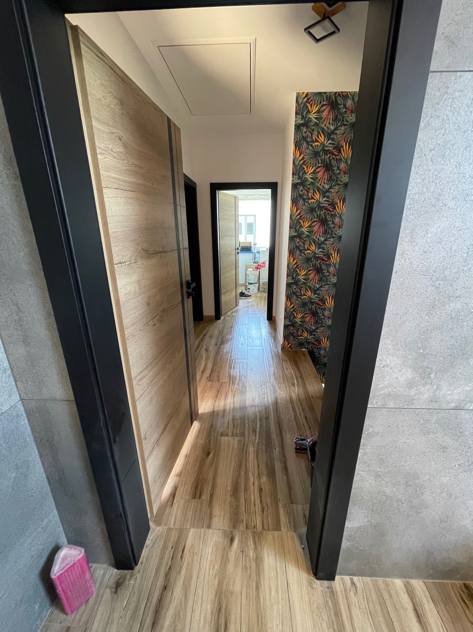 Hall z drewnopodobną podłogą i tropikalną tapetą. Widok przez czarną futrynę, z fragmentem szarej ściany i różowej gąbki w rogu kadru. W tle widoczny fragment kuchni.
