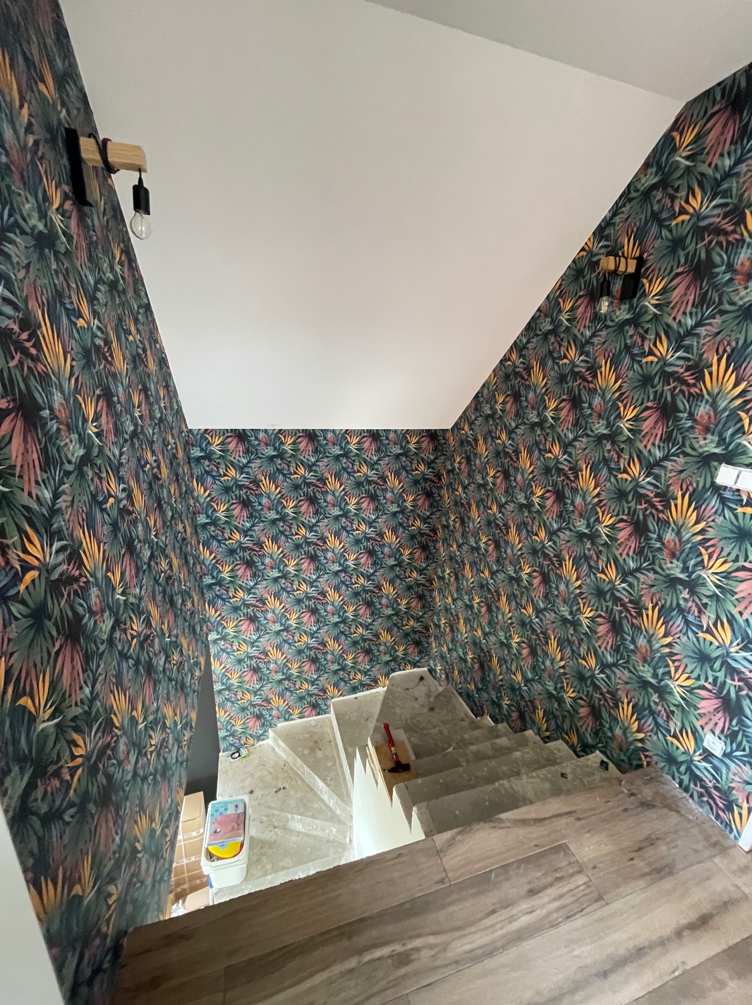 Wnętrze klatki schodowej z wzorzystą tapetą w motywy roślinne, drewnianymi lampami ściennymi i surowymi schodami, częściowo wykończonymi panelami podłogowymi.